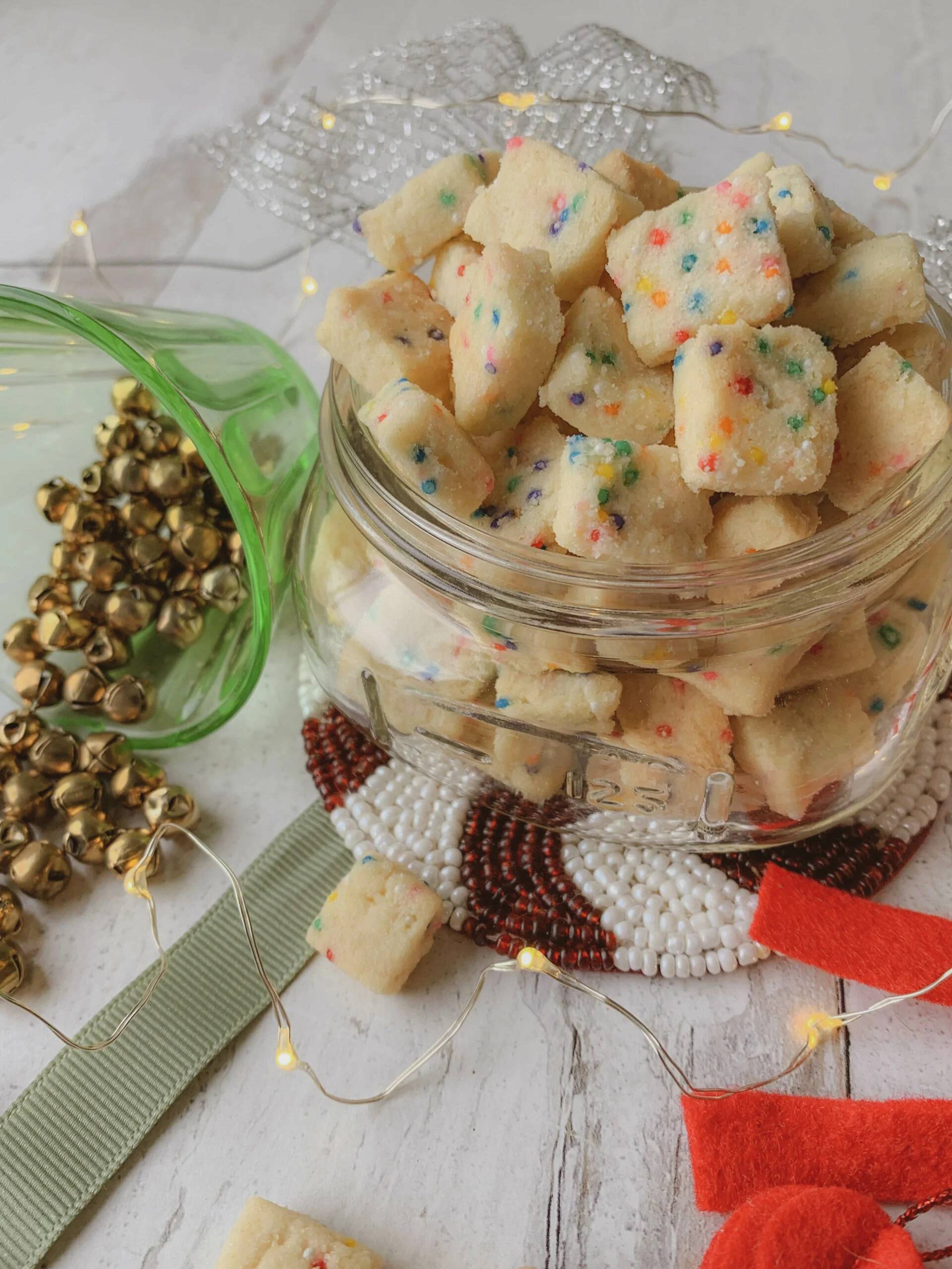 A glass jar filled with small, square, sprinkle-covered shortbread cookies sits on a beaded coaster, surrounded by holiday decorations, jingle bells, and string lights.