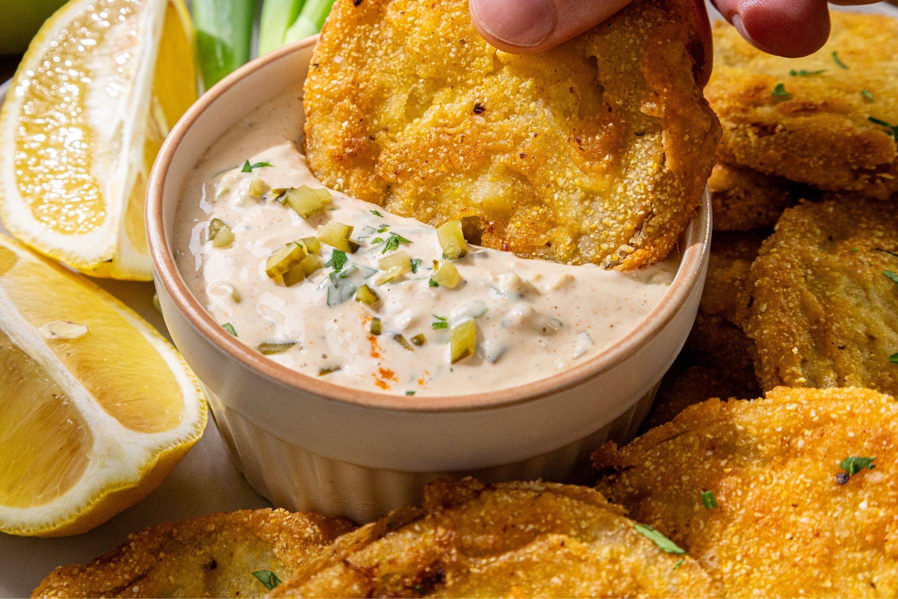 A hand dips a crispy fried green tomato into a creamy sauce topped with chopped pickles and herbs, surrounded by lemon wedges and more fried green tomatoes.