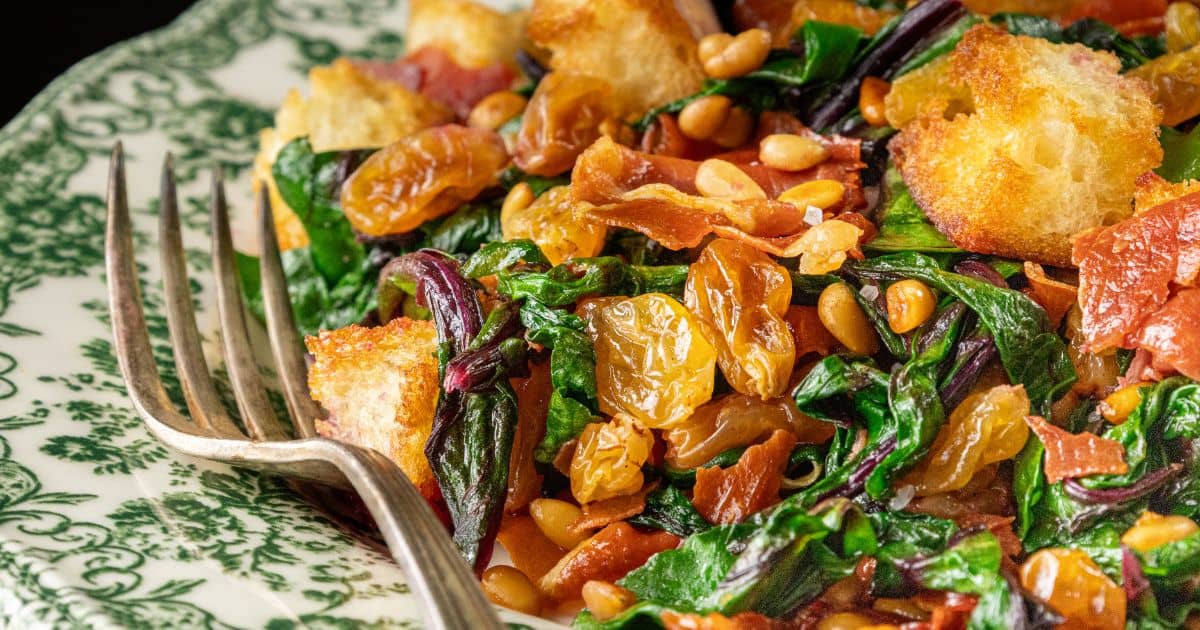 A close-up of a salad with wilted greens, crispy bacon, golden raisins, toasted pine nuts, and crunchy croutons on a decorative green and white plate with a fork beside it.