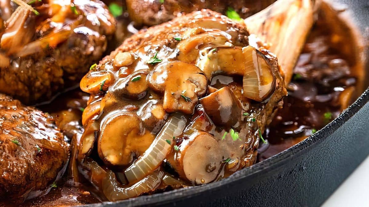 A Salisbury steak in a cast iron skillet with mushroom and onion brown gravy.