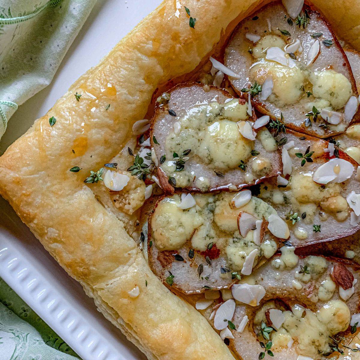 A close-up of a baked puff pastry tart topped with sliced pears, crumbled blue cheese, almond slivers, fresh thyme, and a golden, flaky crust, served on a white plate.