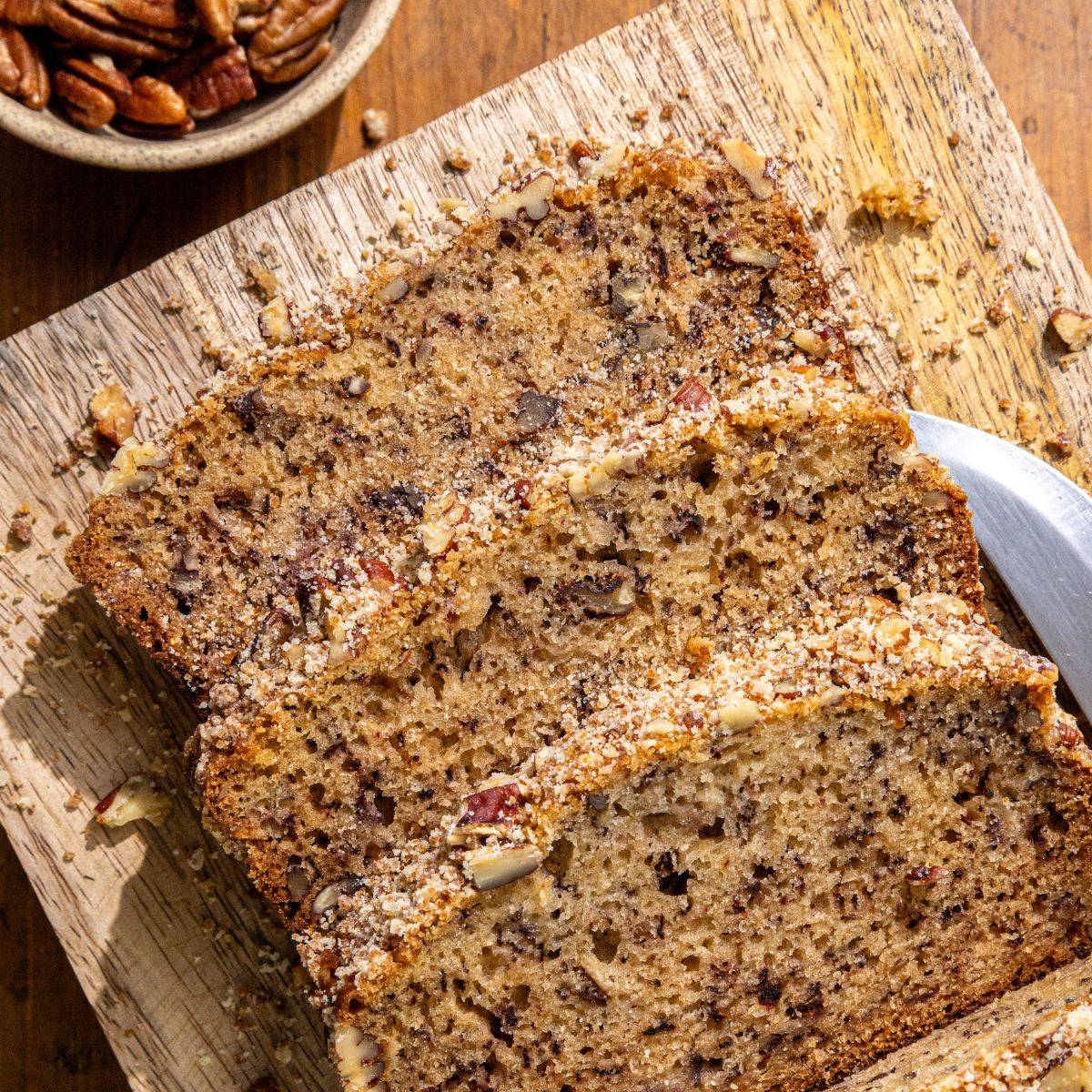 Sliced banana bread with chopped pecans on a wooden cutting board, with a knife beside the loaf and a small bowl of pecans in the background.