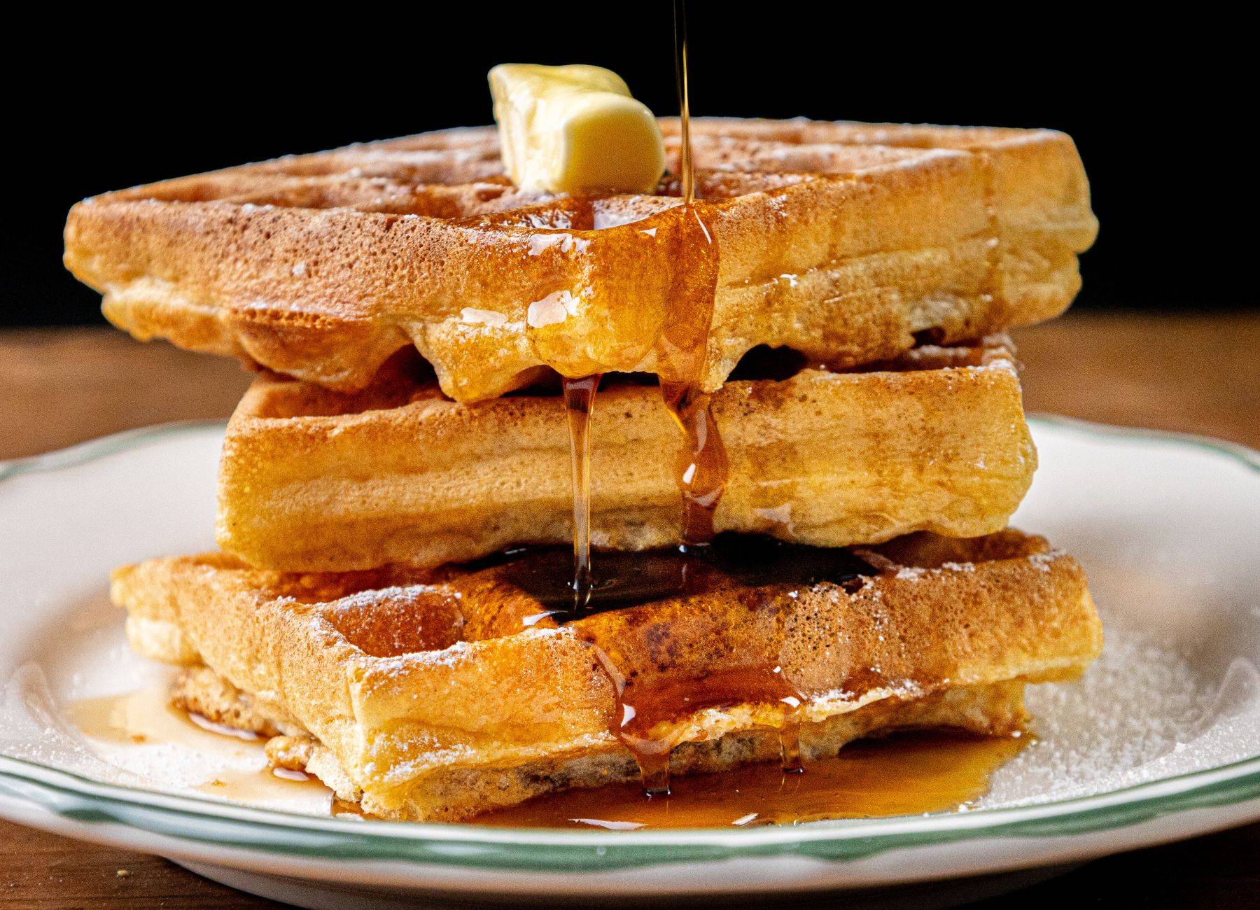 A stack of three golden-brown waffles topped with a pat of butter, with syrup being poured over them. The waffles are on a white plate with a green rim, and the syrup drips down the sides onto the plate. The background is dark, making the waffles and syru