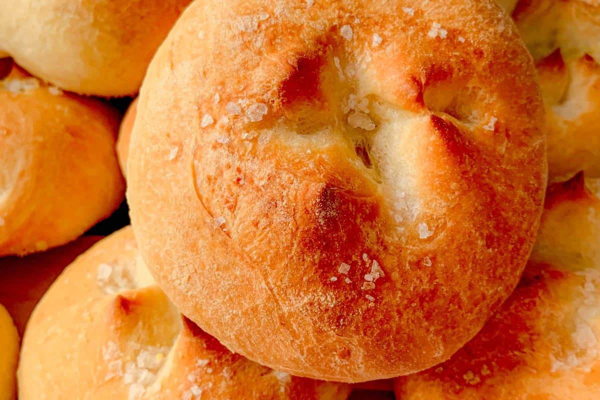 Golden brown, round bread rolls with a crisp crust and a sprinkle of coarse salt on top, stacked closely together.