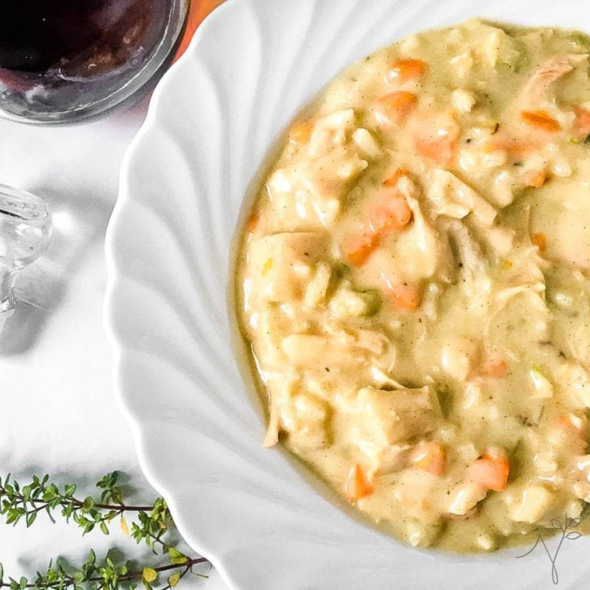 A white plate filled with creamy chicken and vegetable stew sits on a white table near a glass of dark beverage and a sprig of fresh herbs. The stew contains chunks of chicken, carrots, and celery in a thick sauce.