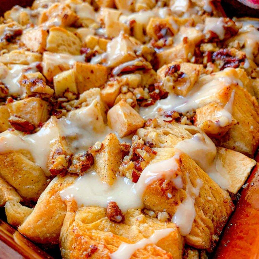 Close-up of a baked dessert with golden biscuit pieces, chopped pecans, and apple chunks, drizzled with creamy white icing in a baking dish.