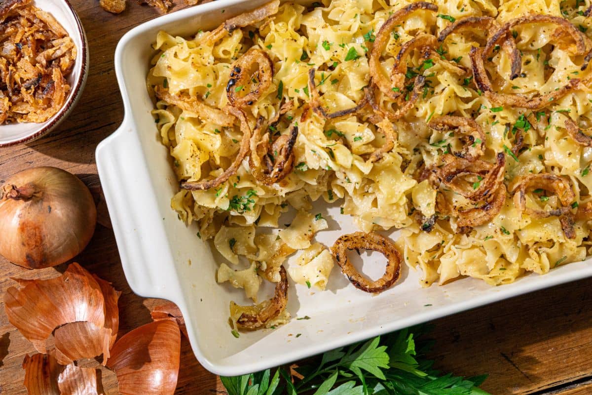 A white baking dish filled with French Onion Noodle Casserole, topped with crispy fried onions and garnished with chopped herbs; onion skins and fresh parsley are nearby on a wooden surface.
