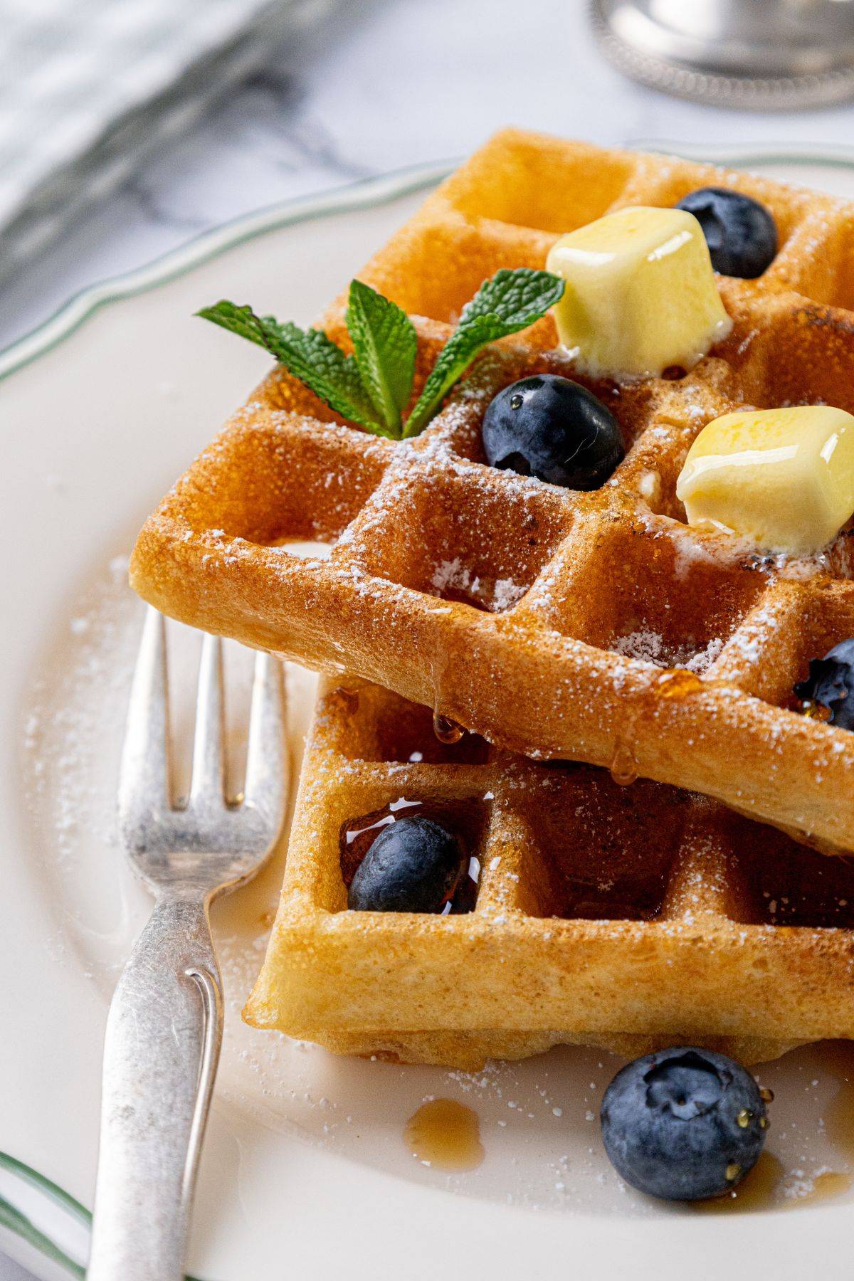 A close-up of two stacked waffles on a plate, garnished with blueberries, a sprig of mint, and two pats of butter. The waffles are lightly dusted with powdered sugar and drizzled with syrup. A fork is placed beside the waffles.