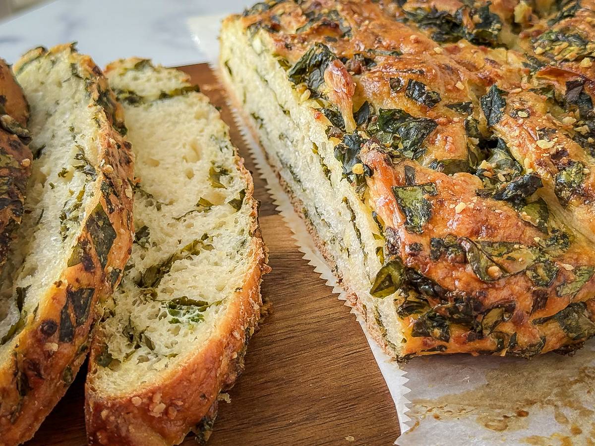 Sliced homemade bread with a golden crust topped and filled with chopped green herbs, resting on a wooden cutting board with parchment paper underneath.