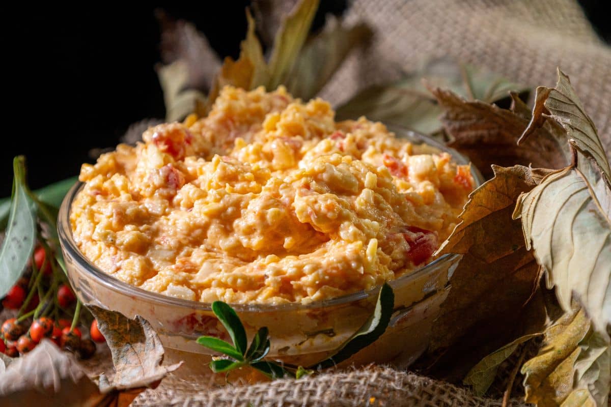 A glass bowl filled with creamy pimento cheese dip sits surrounded by autumn leaves and berries on a rustic burlap surface.