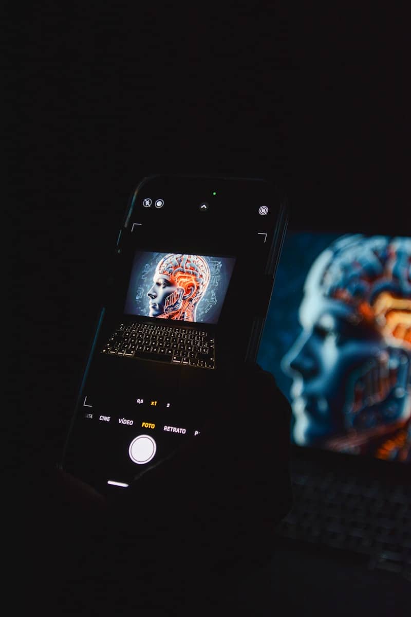  A dark photo of a smartphone held in front of a laptop. The phone screen shows an image of a human head with an illuminated brain diagram over a keyboard, creating a moody, tech-inspired composition. The laptop screen in the background displays a similar