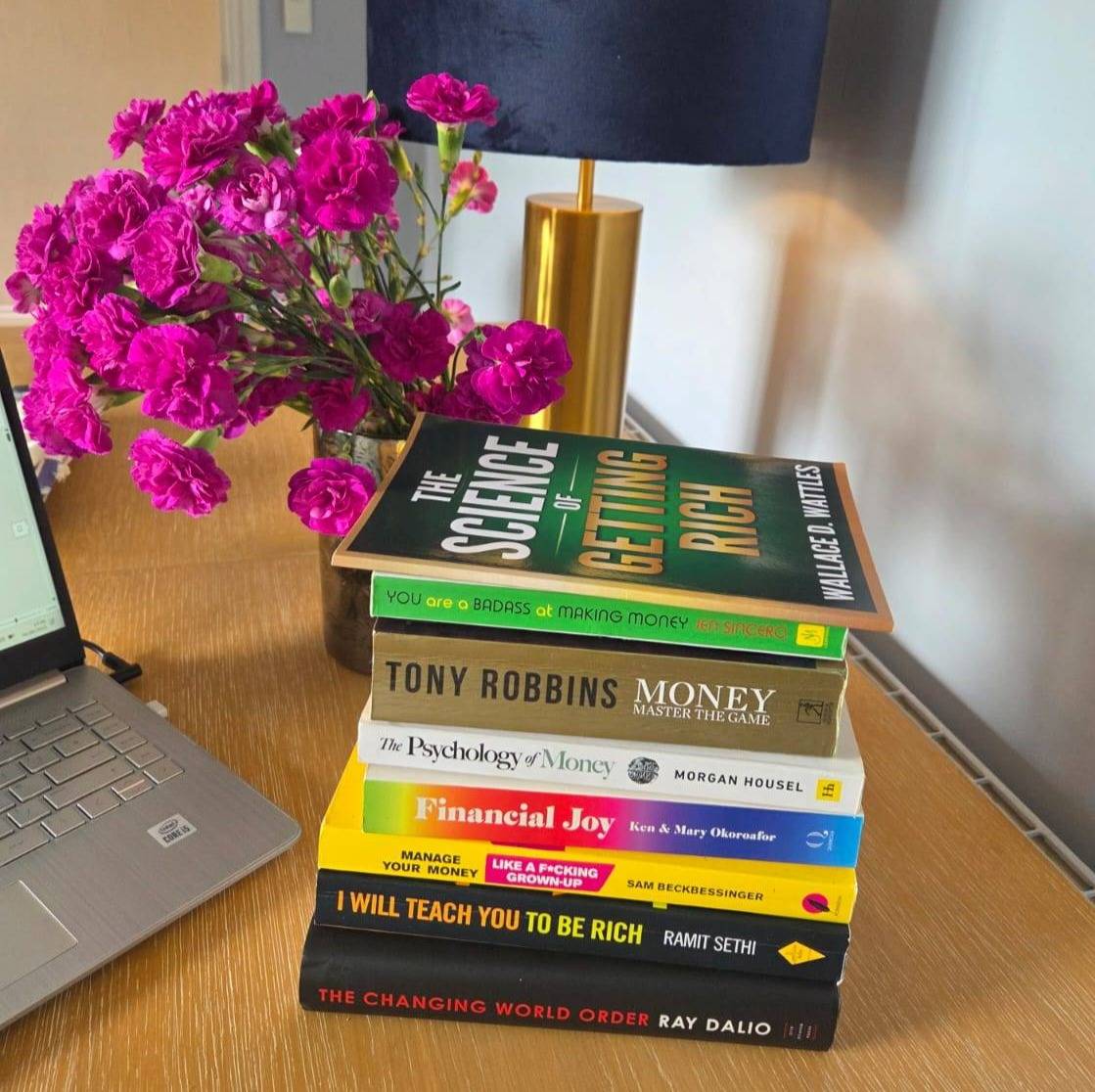 A laptop and bright pink flowers in the background, up front a pile of money and finance books.