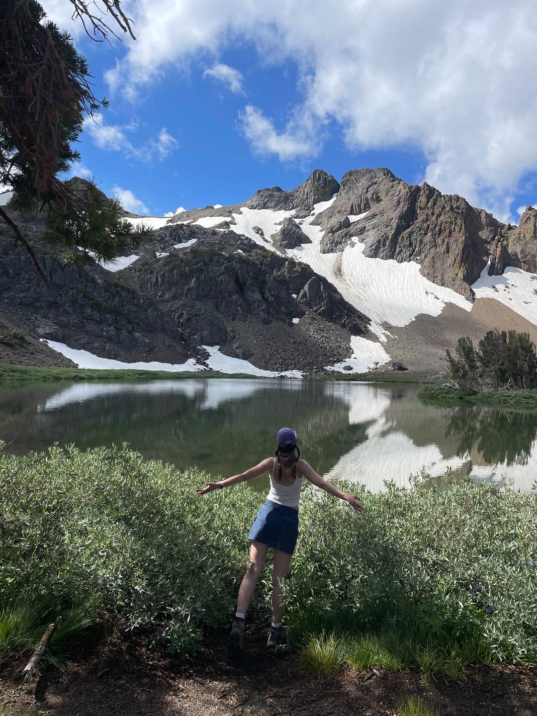 Hanna with arms flung outward while taking in the view at Round Top Lake in California. The mountain peaks and lingering snow drifts are reflected in the water.
