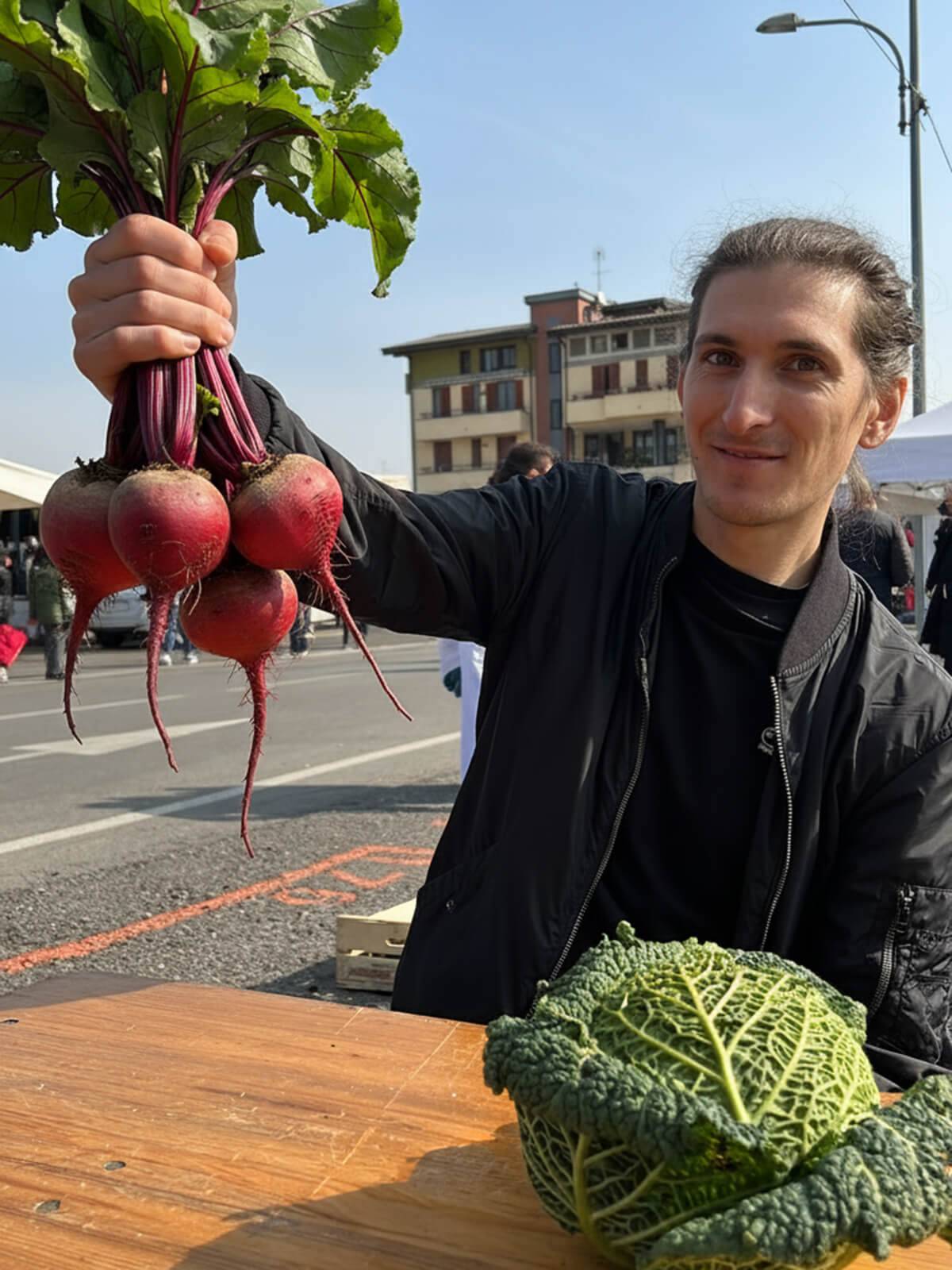 Persona joven sosteniendo varias remolachas frescas con raíces largas, en un mercado al aire libre.