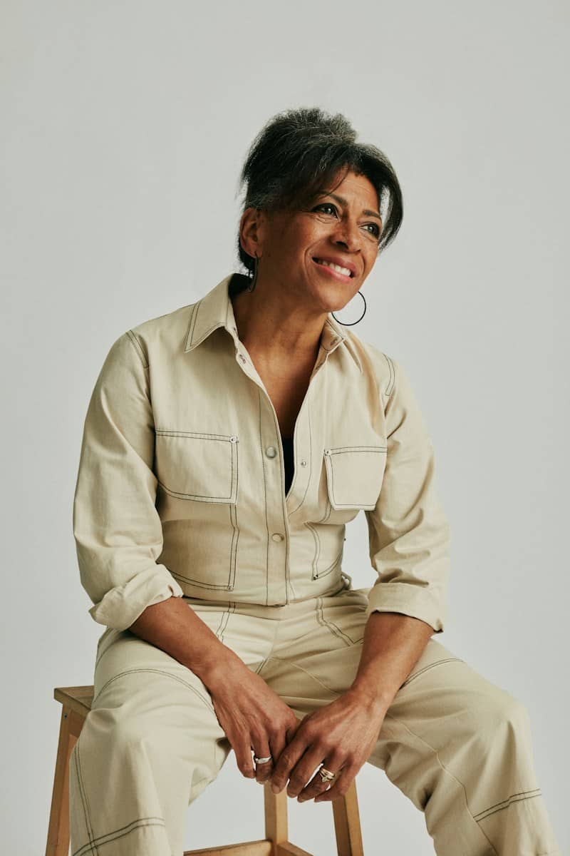 Woman in beige jumpsuit smiling, sitting on a stool