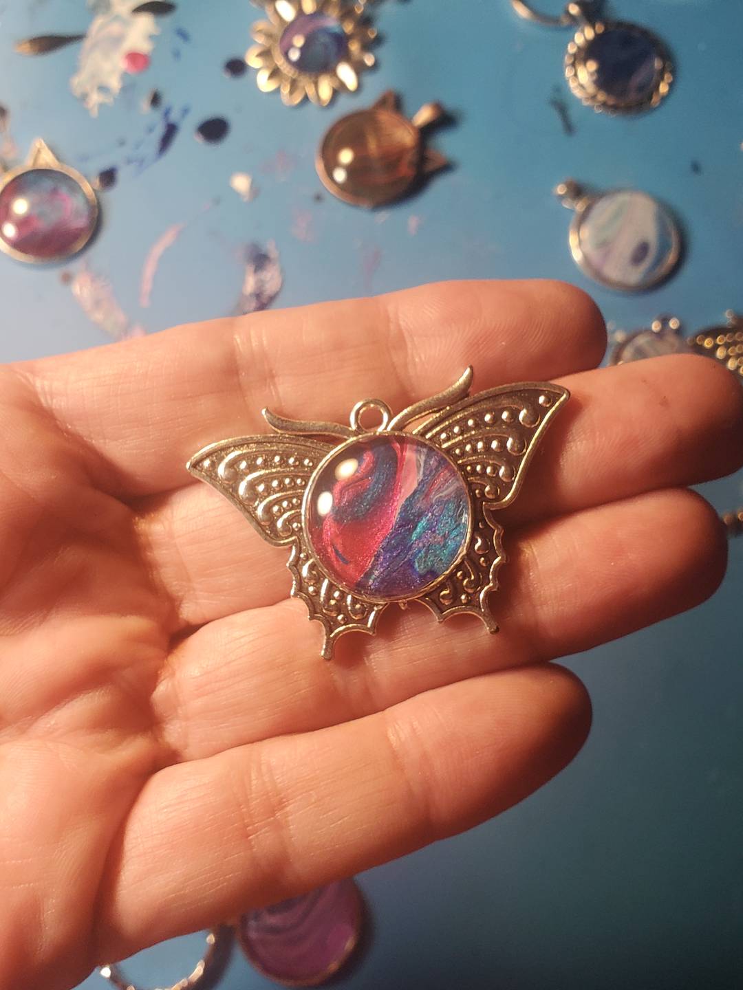 close up of a hand holding a butterfly pendant with colors of blue and pink inside - behind the hand there are other pendants laying on a table.