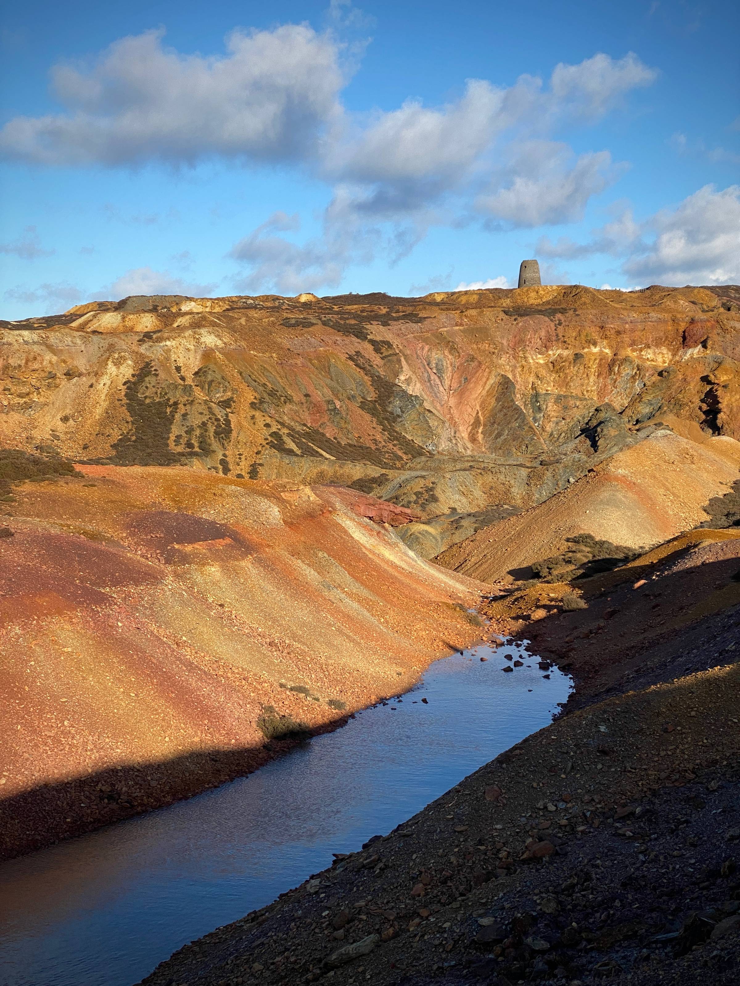 A colourful red and yellow open cast mine with a ruined windmill on the horizon, blue water in foreground and blue sky with grey clouds.