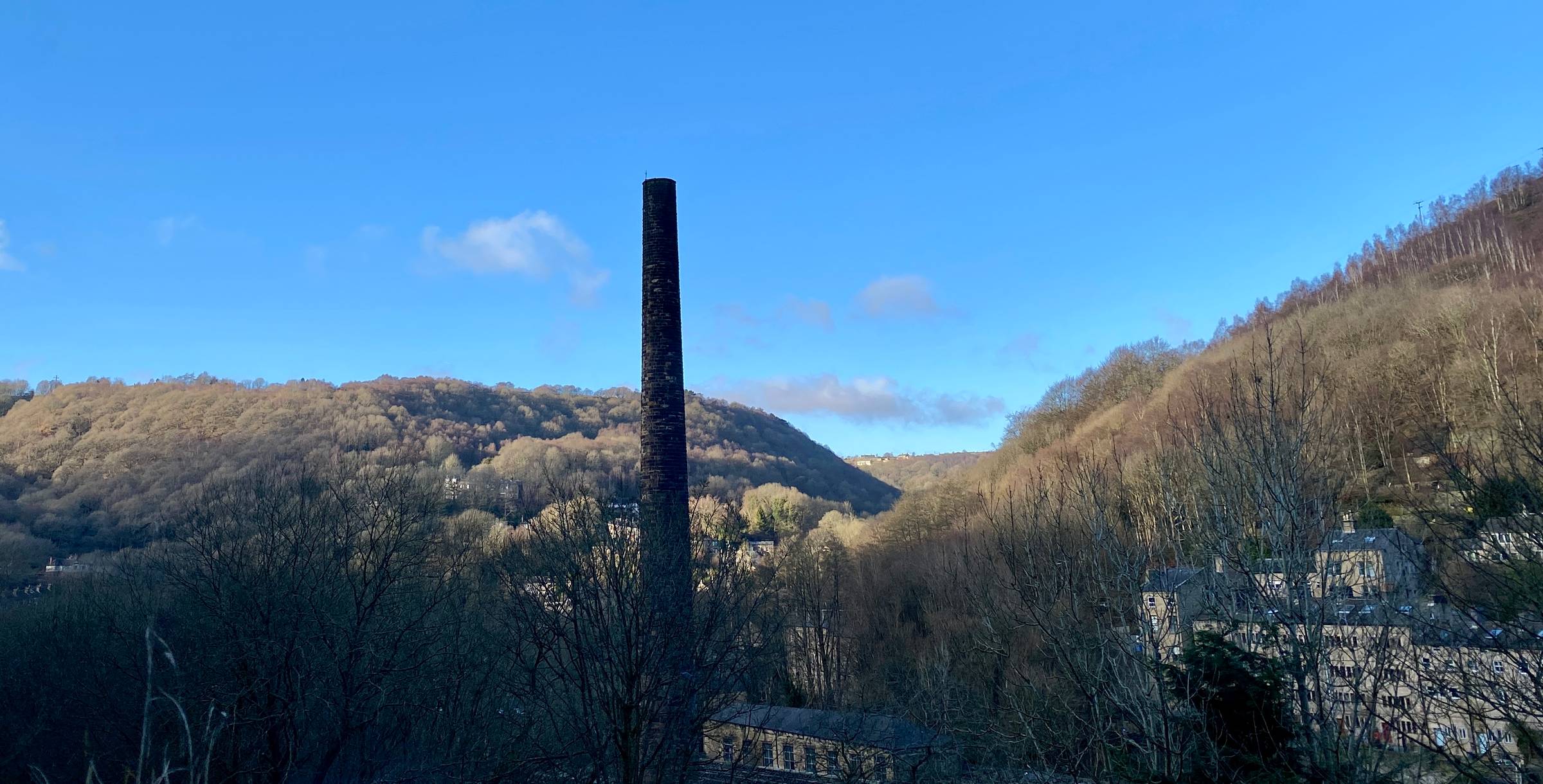 A tall dark mill chimney rising from woodland into a pure blue sky.