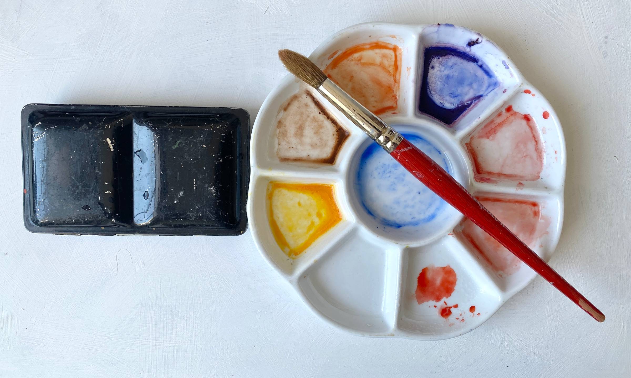 A circular porcelain paint palette with watercolour paints in use and red paintbrush.