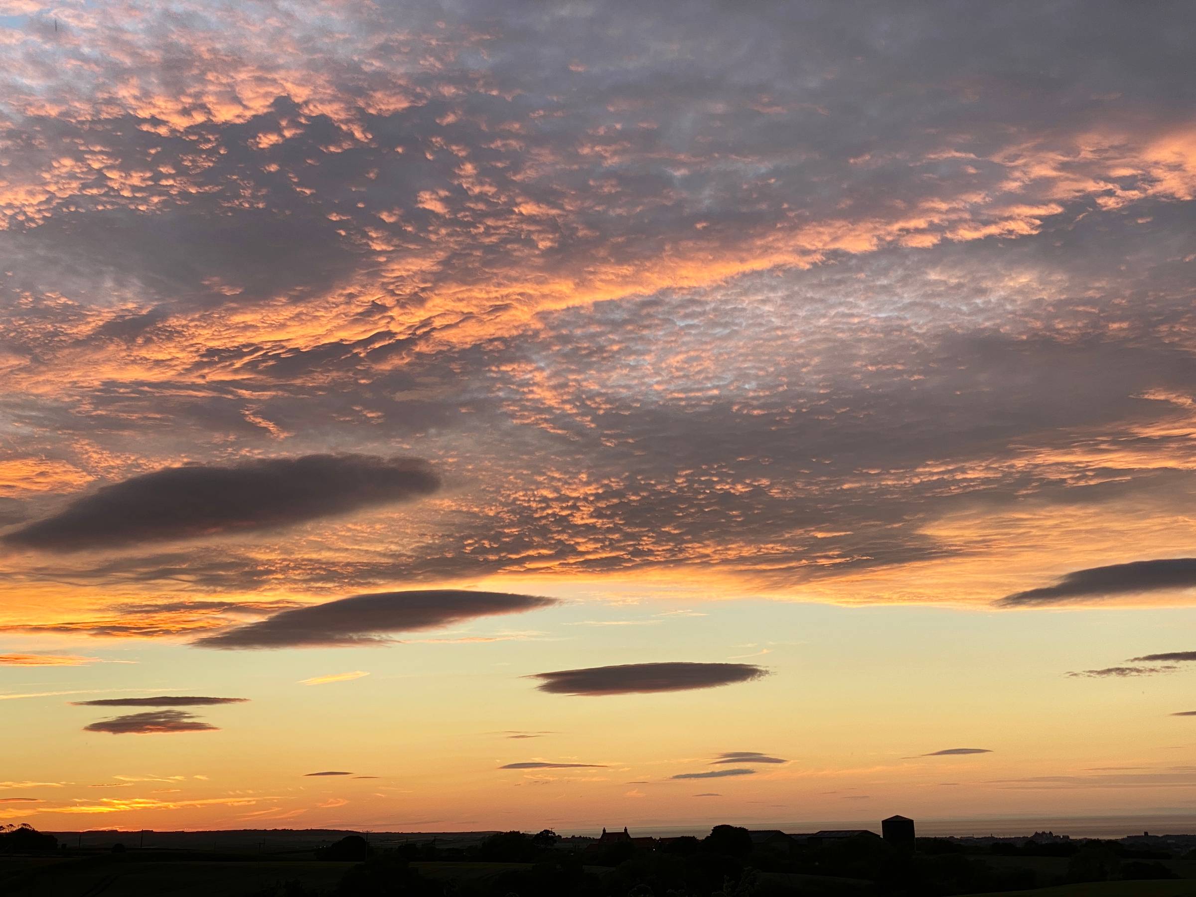 A sunset over the sea coast with very dark land and pinky golden light with complex clouds extending high up.