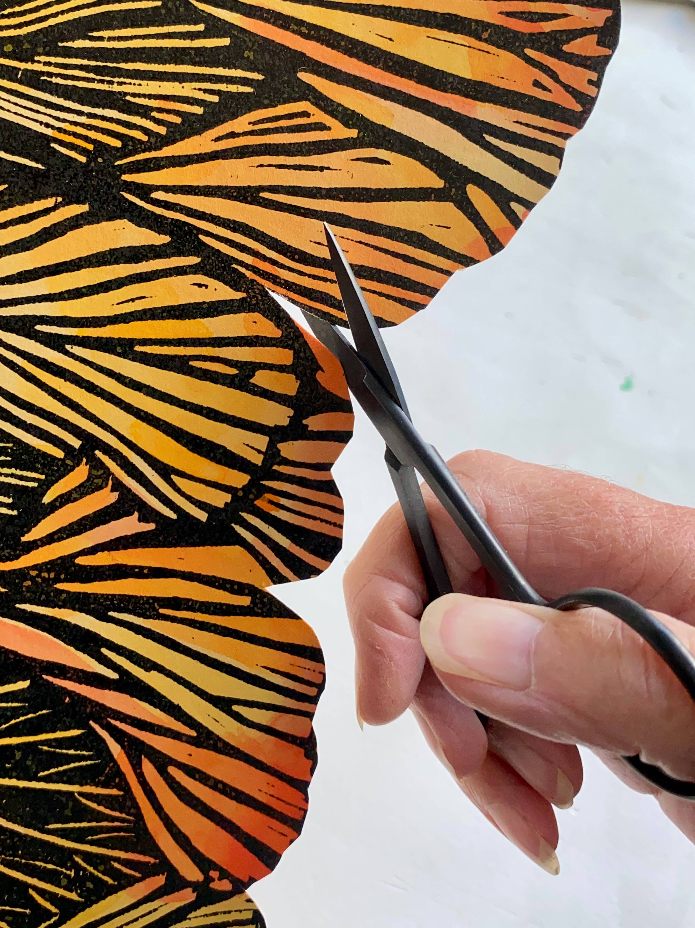 A person's hand with small scissors cutting out an artwork with orange and lack texture.