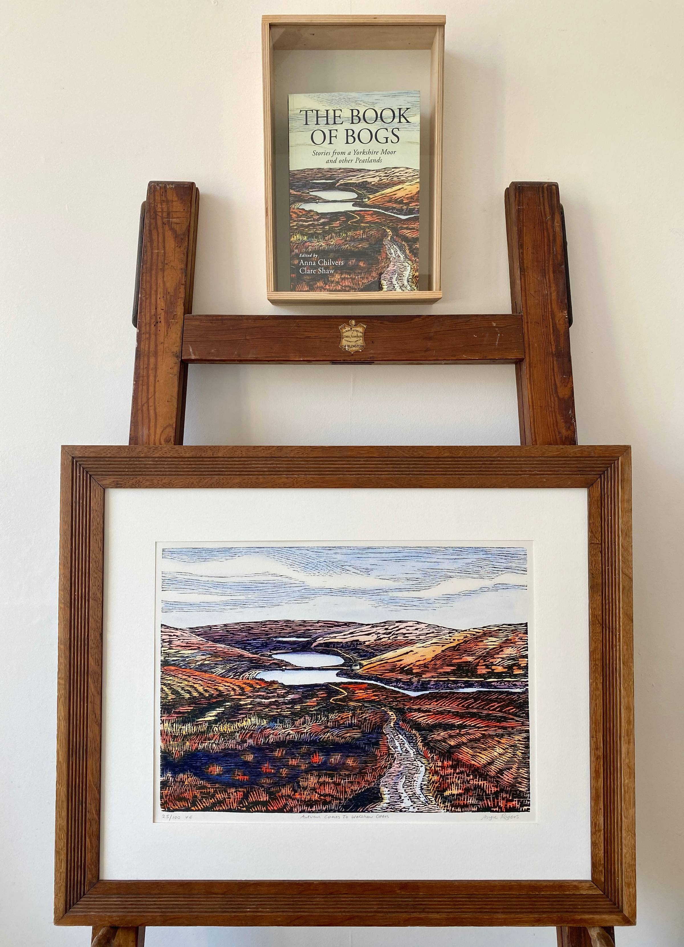 An old wooden easel and frame with a woodcut artwork of autumnal moorland in red and orange tones.