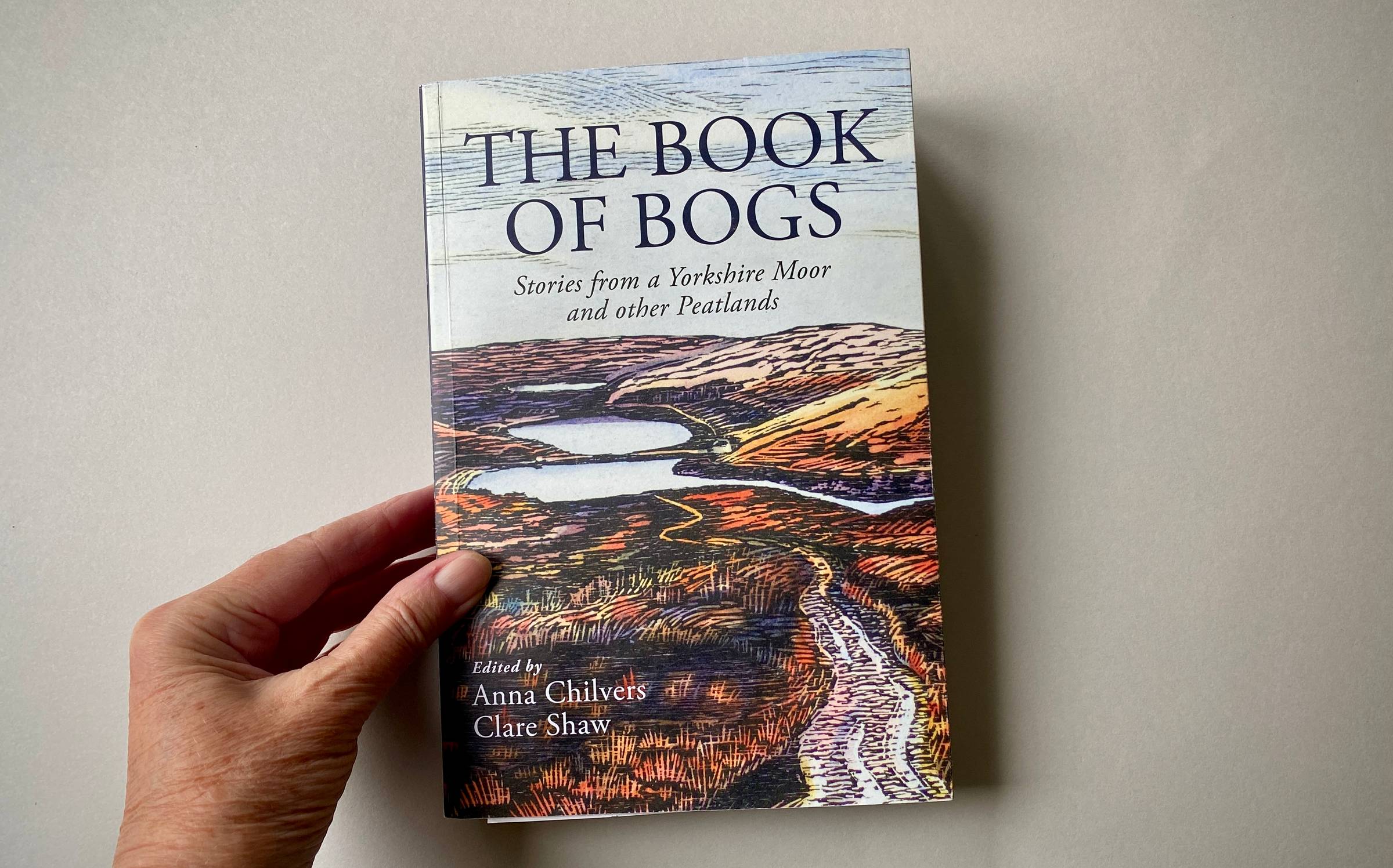 Hand holding large paperback book with autumn woodcut print landscape on cover.