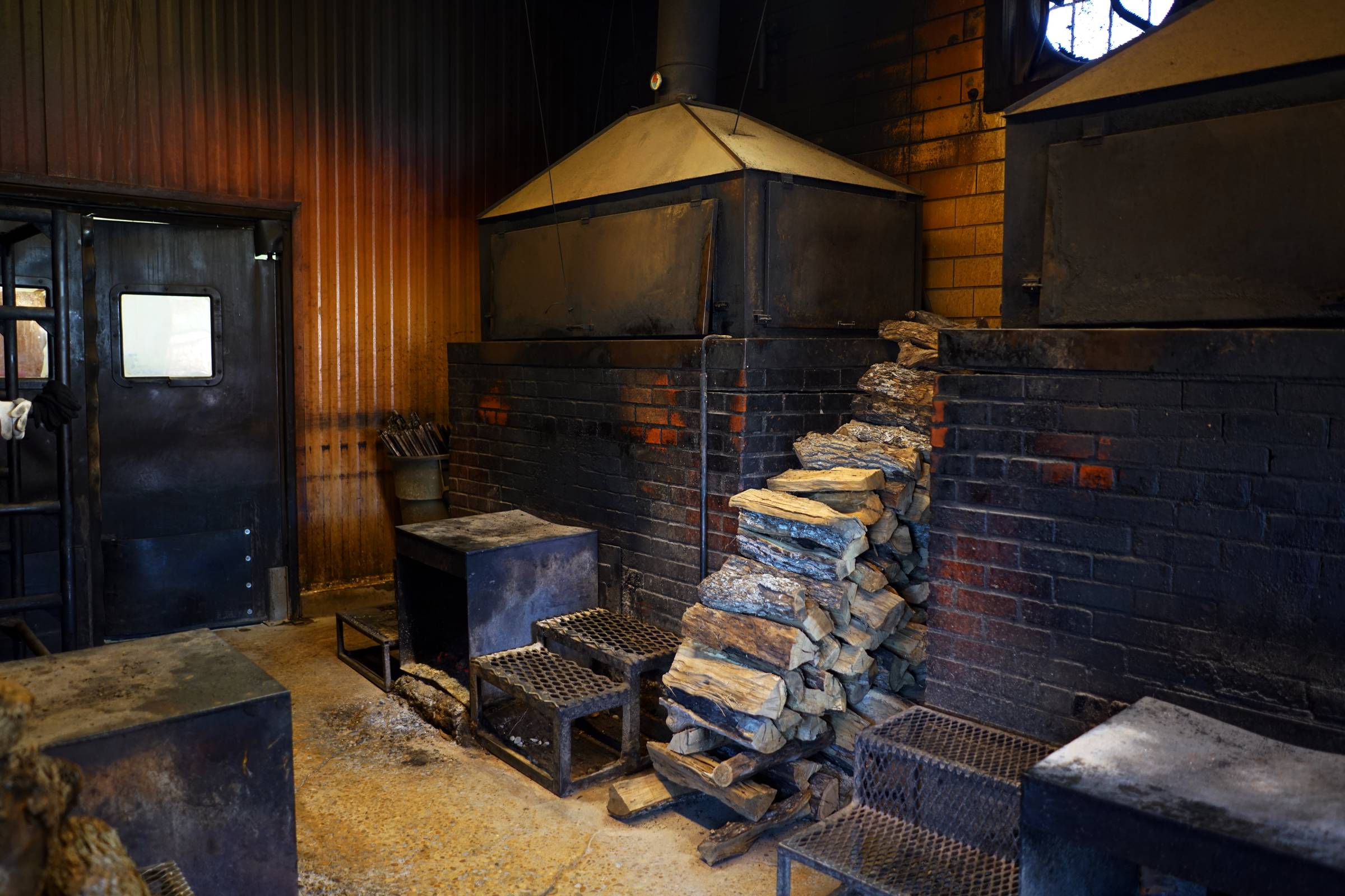 Two brick smoker units inside a dingy room with a pile of firewood stacked between them.