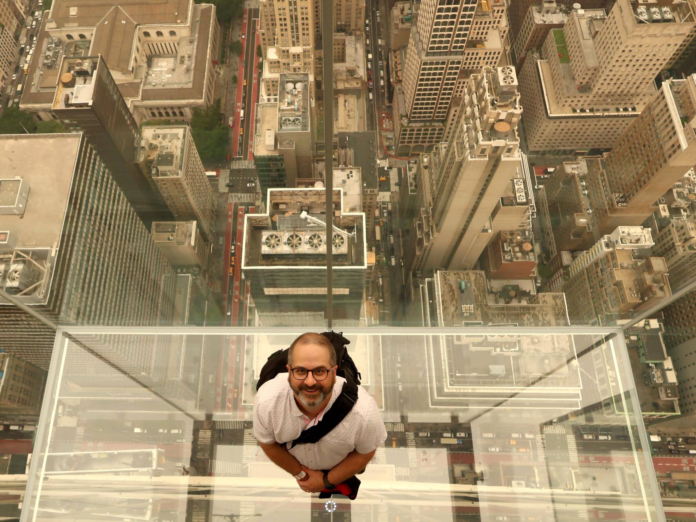 Photo looking down at man on glass-floored observation deck far above other skyscrapers