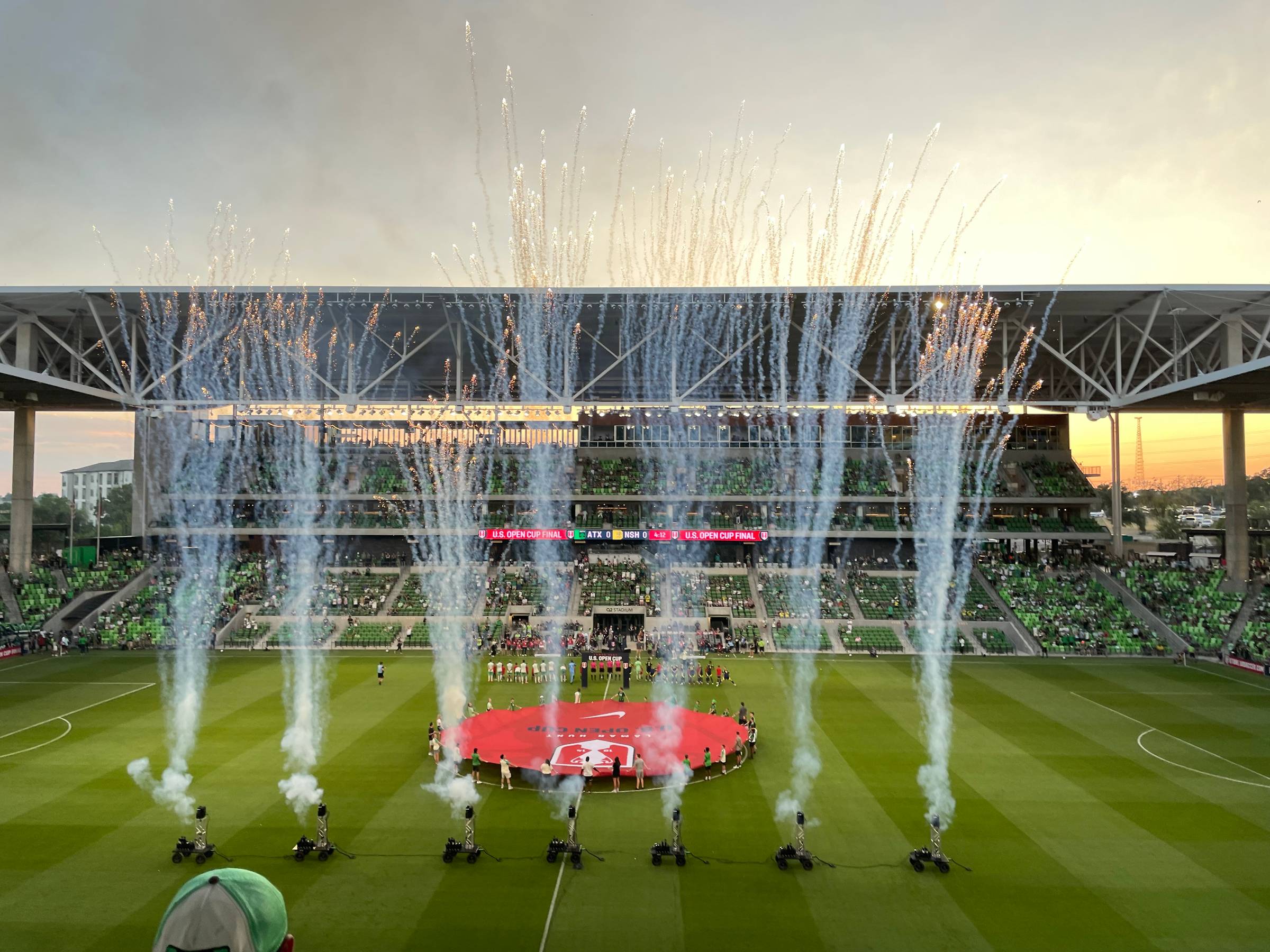 7 columns of fireworks and colored smoke shoot up from the soccer pitch inside a soccer stadium.