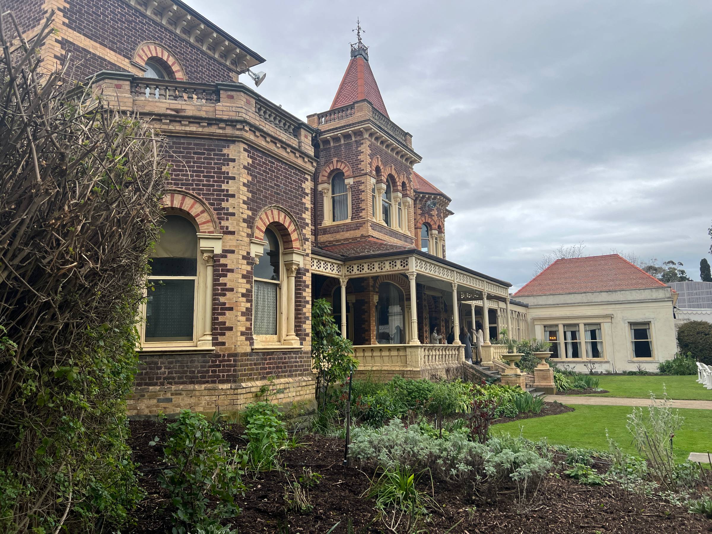 Exterior view of Rippon Lea Estate in Melbourne, showing the ornate Victorian mansion with its dark brickwork, cream detailing, tower and red-tiled roof, surrounded by landscaped garden beds and lawns beneath an overcast sky.