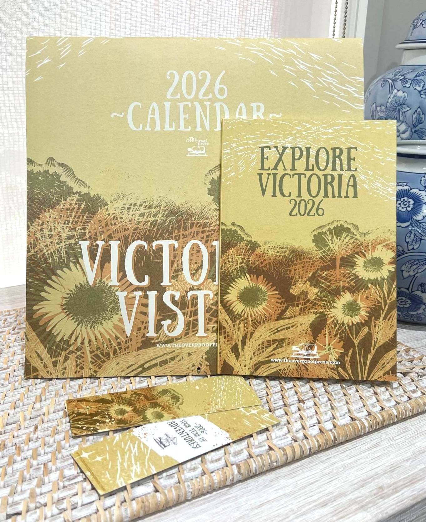 Flatlay of the Overproof Press 2026 Victorian Vistas Calendar and Explore Victoria Zine on a wooden table, surrounded by interior decor including blue vases and mat, photographed in soft natural light.