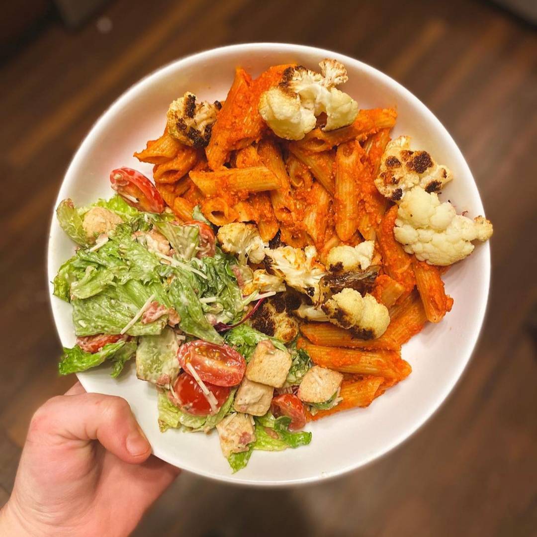 White hand holding a bowl of red pepper pasta with salad on the side