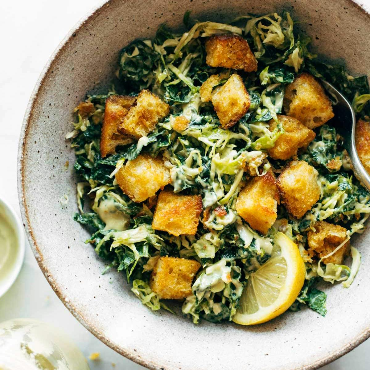 Kale and Brussels Caesar Salad in a bowl with cheezy garlic croutons and a lemon slice