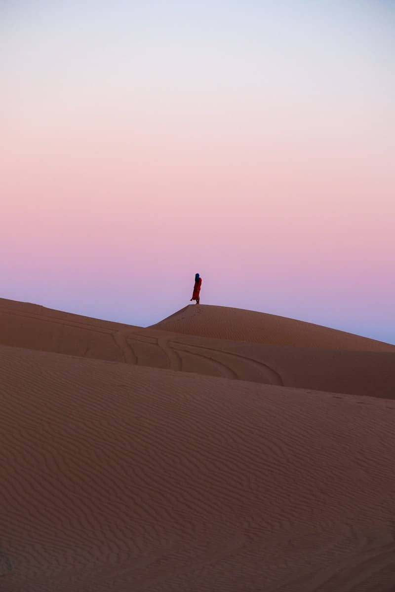 A person stands on a desert dune at sunset.