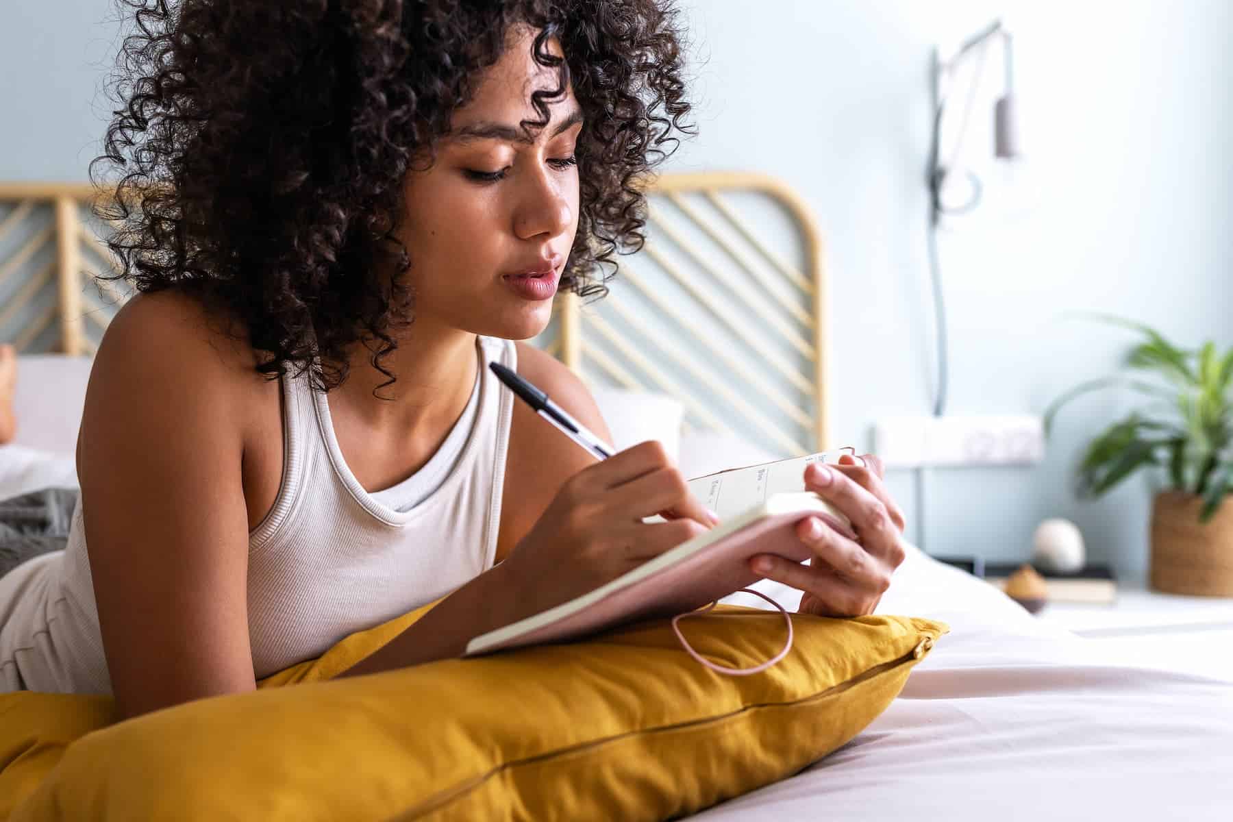 A woman with curly hair lies on her bed, writing in her notebook as she works through her self-care checklist.