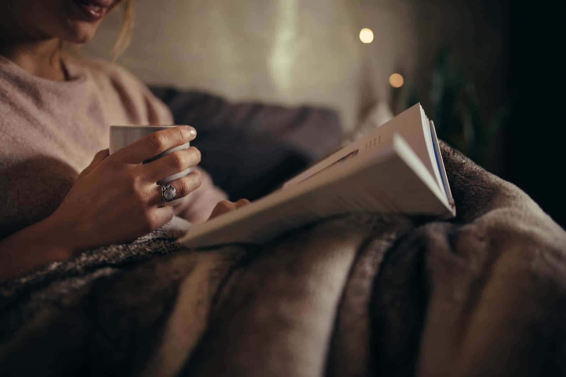 Woman enjoying a quiet evening on the sofa with a hot drink and a book.