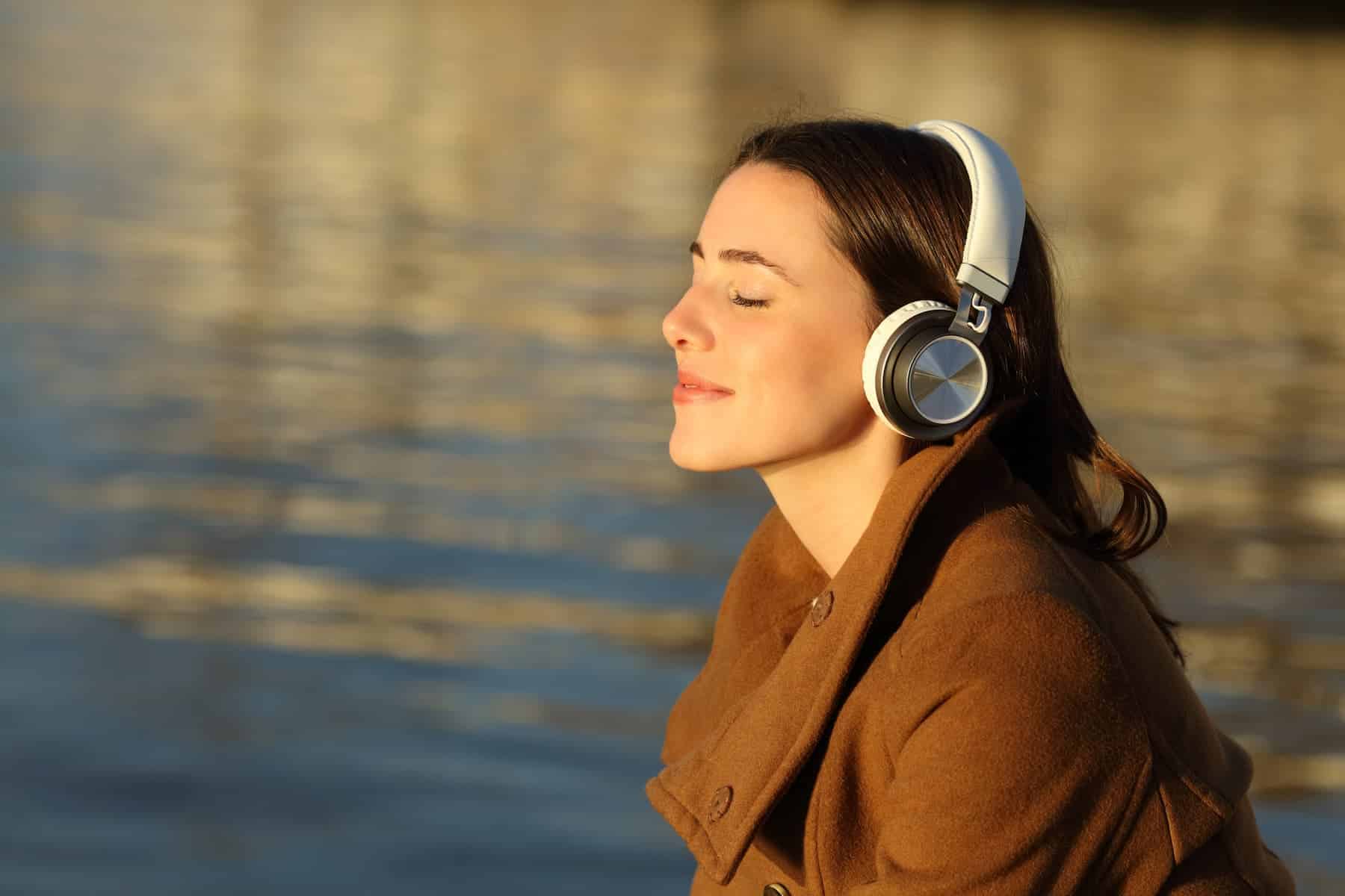 A woman in headphones and a brown coat stands by water with eyes closed, appearing relaxed and content.