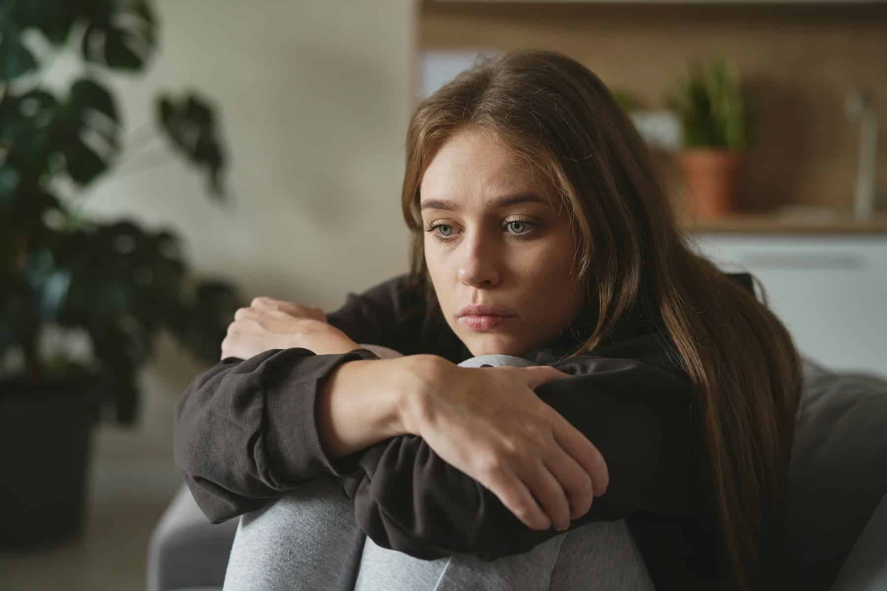 A woman with long brown hair sits curled up with knees to her chest, arms wrapped around herself, looking away sadly.