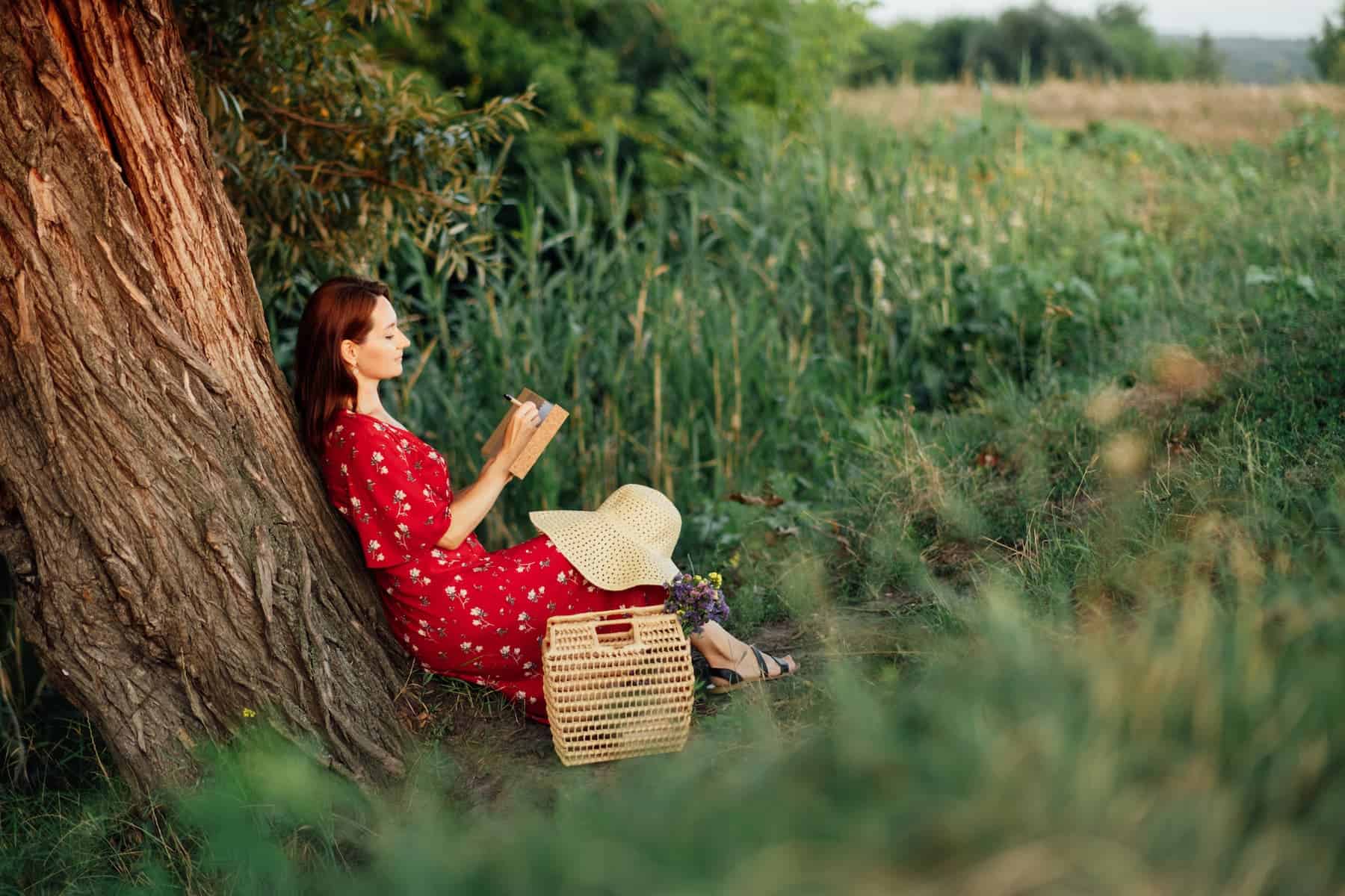 A woman in a red dress relaxes under a tree while reading a book, enjoying a peaceful self care Sunday moment outdoors.