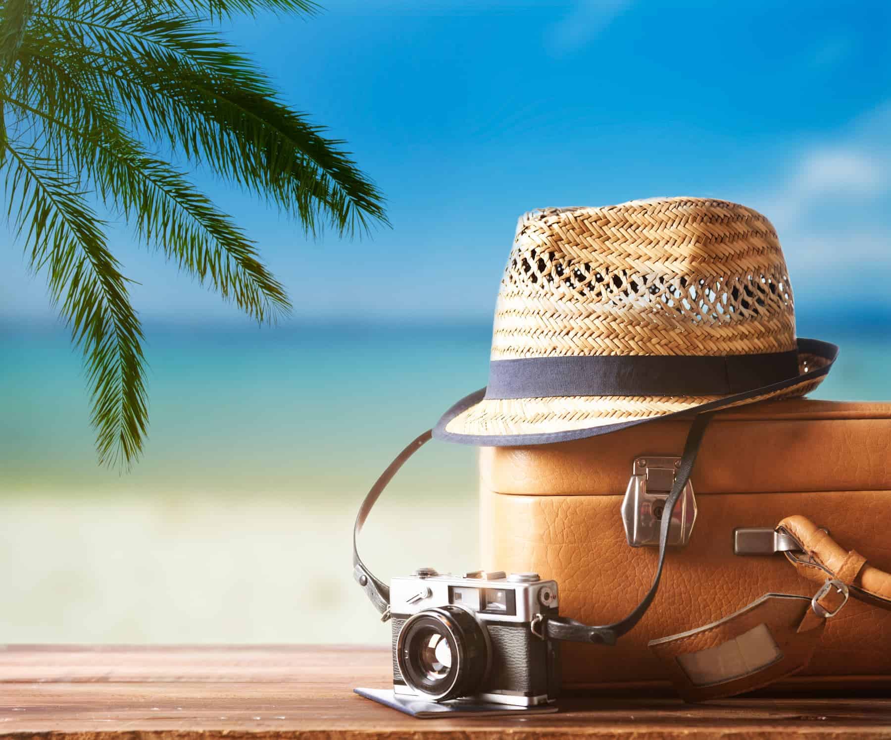 A vintage suitcase, hipster hat, camera, and passport rest on a wooden deck with the beach in the background — a perfect scene that captures how to make packing less stressful.