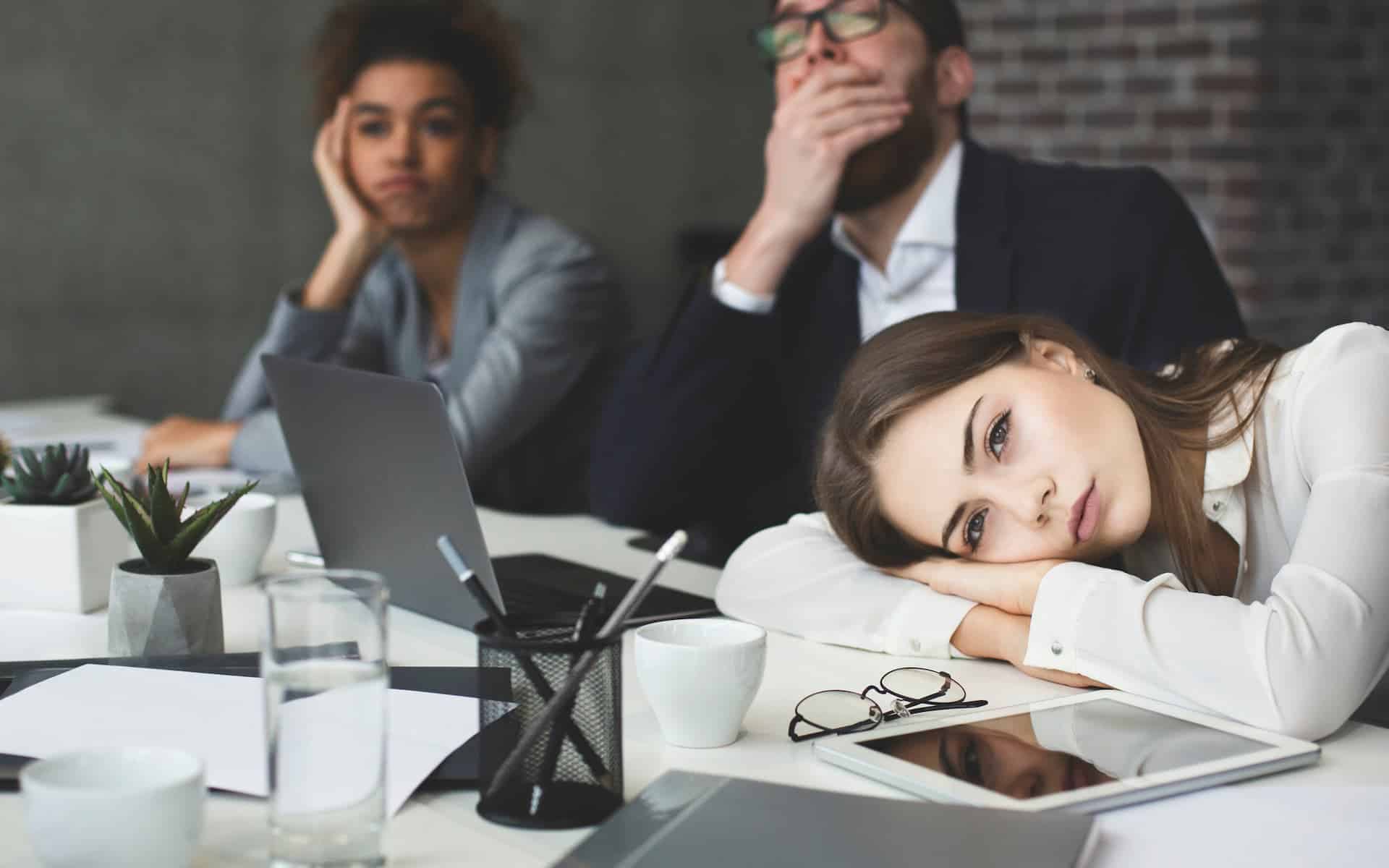 Three people sit at a table looking bored—one rests her head on her arms, one yawns, and one leans on her hand.