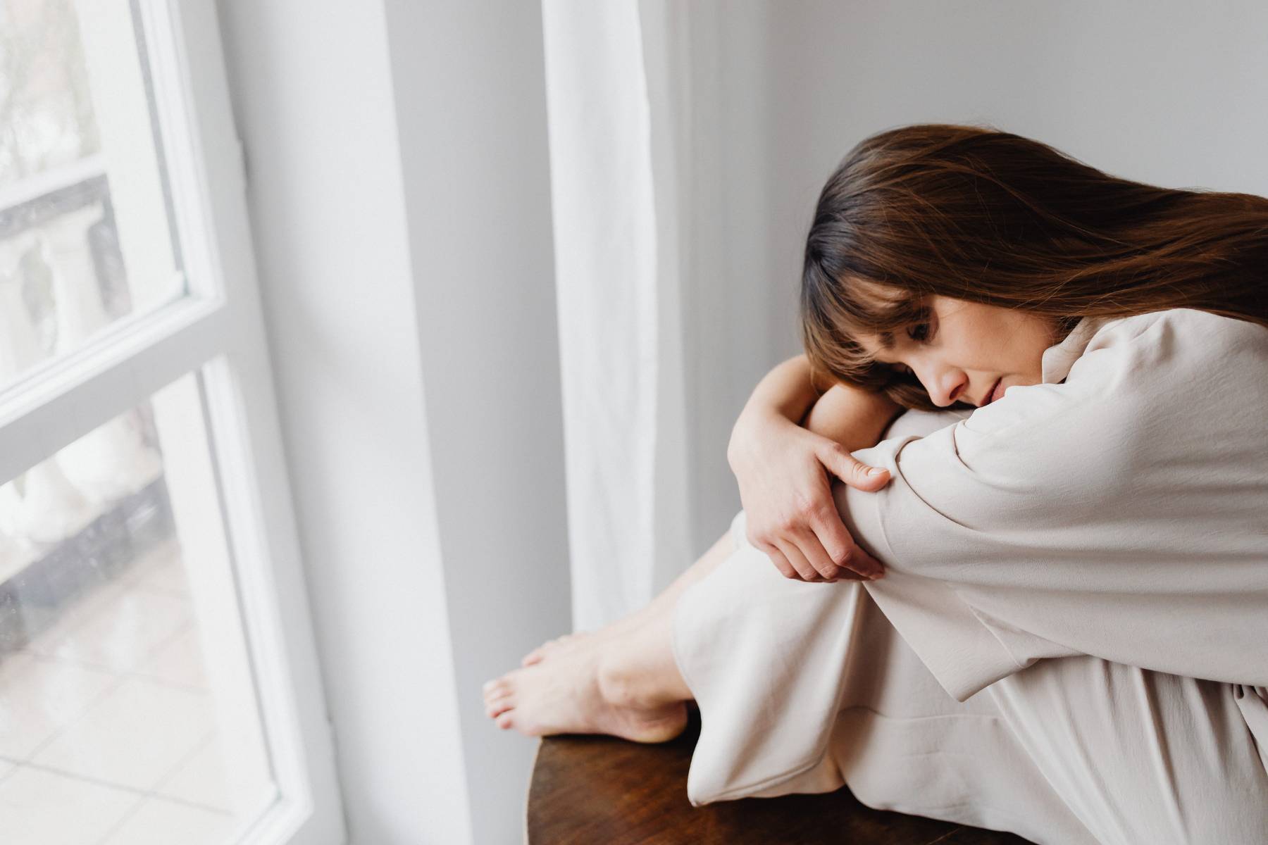 Woman sitting by a window with her knees drawn up, lost in thought.