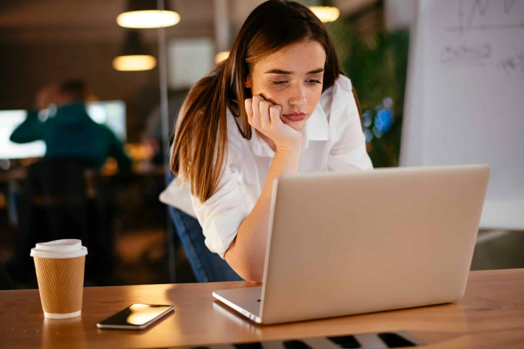 A woman leans on a desk and stares at a laptop with a coffee cup and phone nearby, appearing bored at her office job.