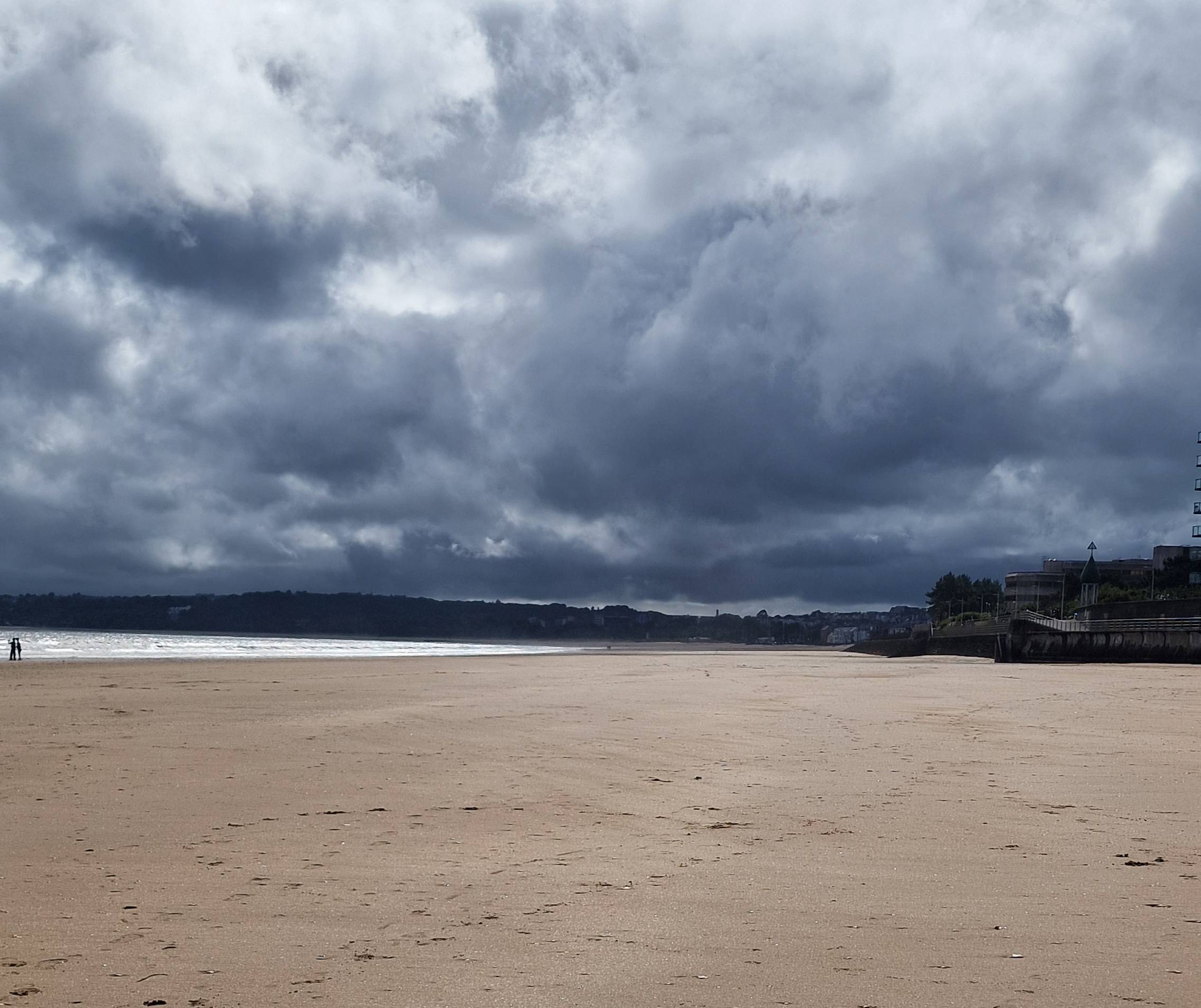 Wide sandy beach at Swansea Bay beneath dramatic summer clouds, with sunlight breaking through over the sea and a peaceful, open shoreline.