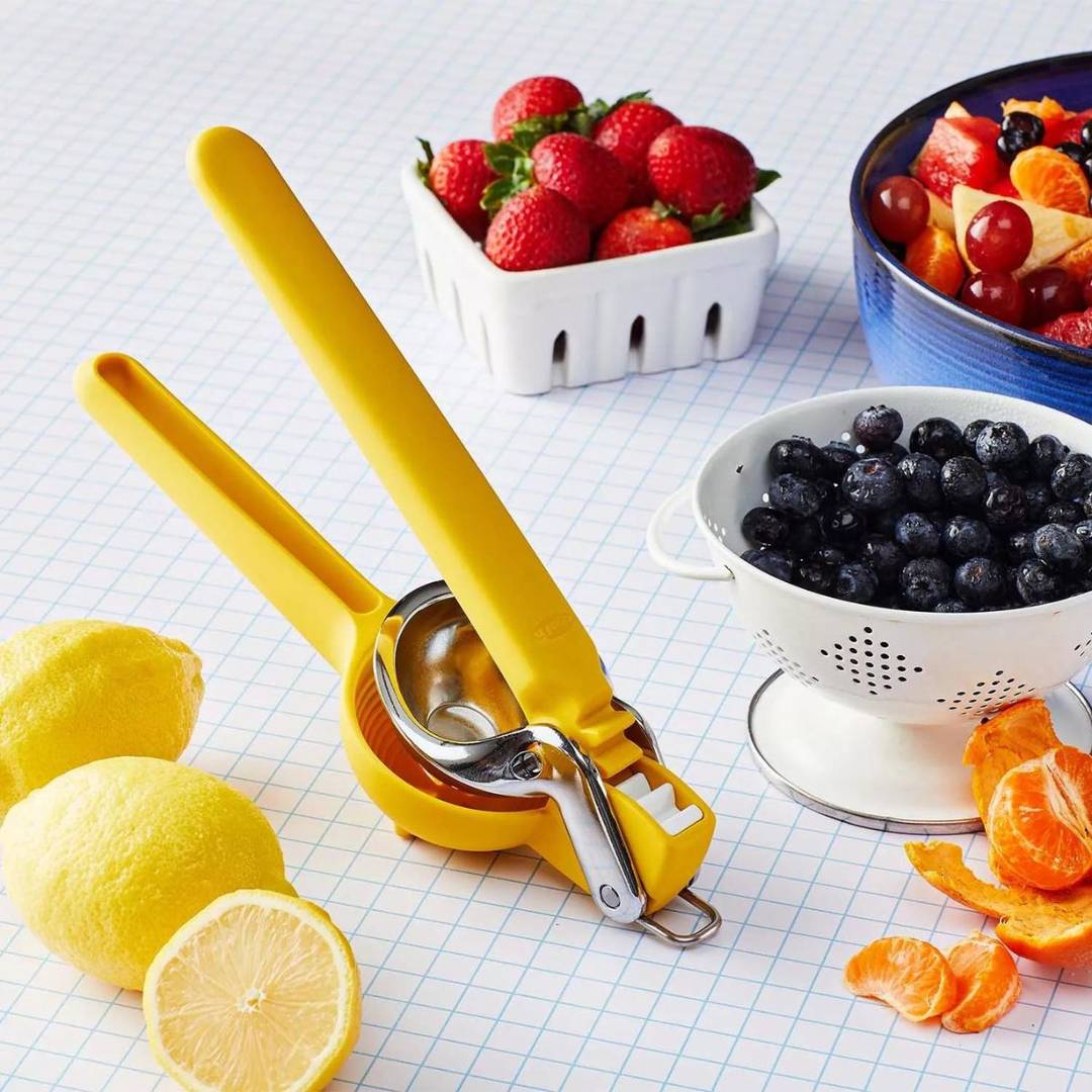 A yellow citrus juicer, hand held on a counter with berries, citrus around it. 