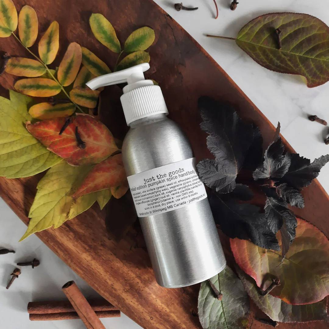 an aluminum bottle with a pump has a label that reads "just the Goods limited edition pumpkin spice hand/body lotion". The rest of the label is too small to read, but it contains the ingredients of the product. The bottle is laying flat in a long oval wooden bowl resting diagonally across the image from upper left to bottom right. In the bowl, below and adjacent to the skin care products is an assortment of autum leaves. Sprinkled on the leaves and onto the marble tile surface under/around the bowl are dried cloves, two cinnamon sticks, and more autumn leaves