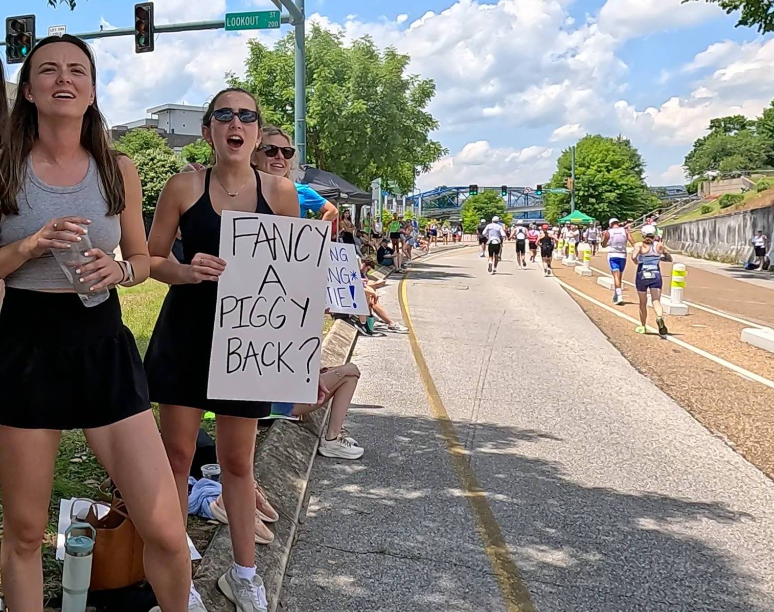 Female spectator holding an encouraging sign that says "Fancy a piggy back?" as triathletes run in the background