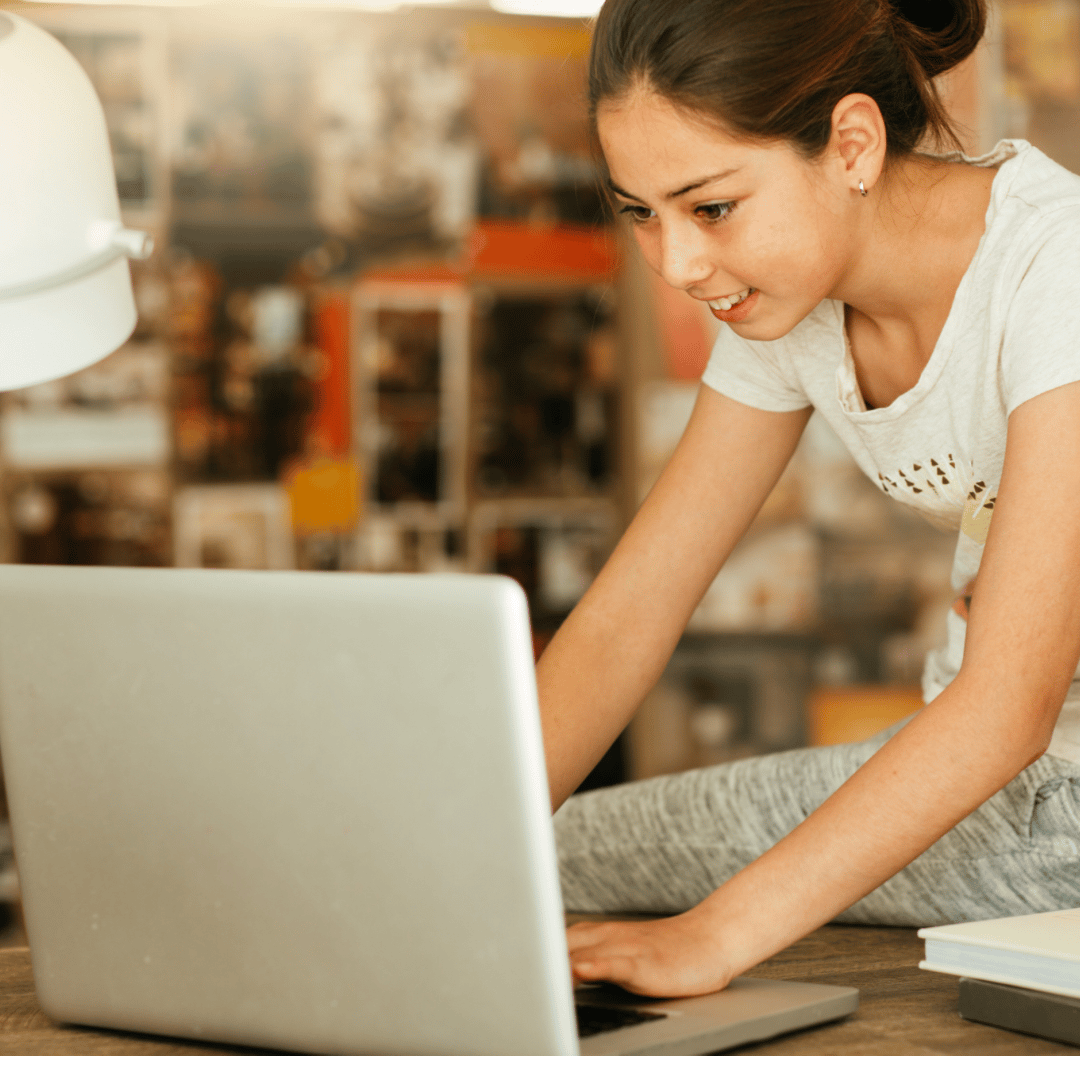 A young person is leaning over a table, using a laptop. They have their hair pulled back and are wearing a casual T-shirt. The background is blurred, showing a colorful interior that appears to be an office or a creative workspace. A white desk lamp is visible on the left side.