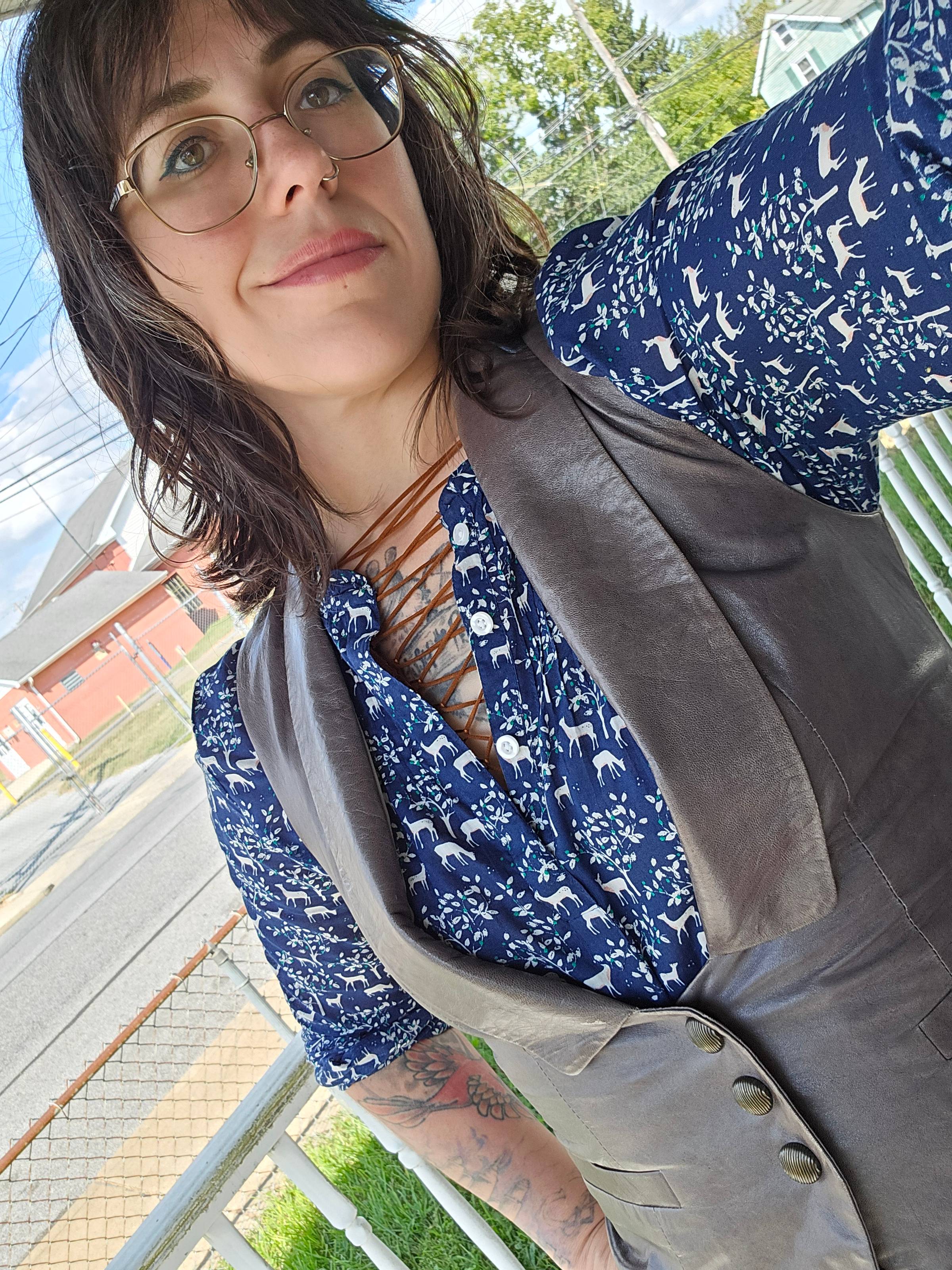 Woman with medium length brown hair and glasses smirking in a gray leather vest and blue deer printed shirt. 